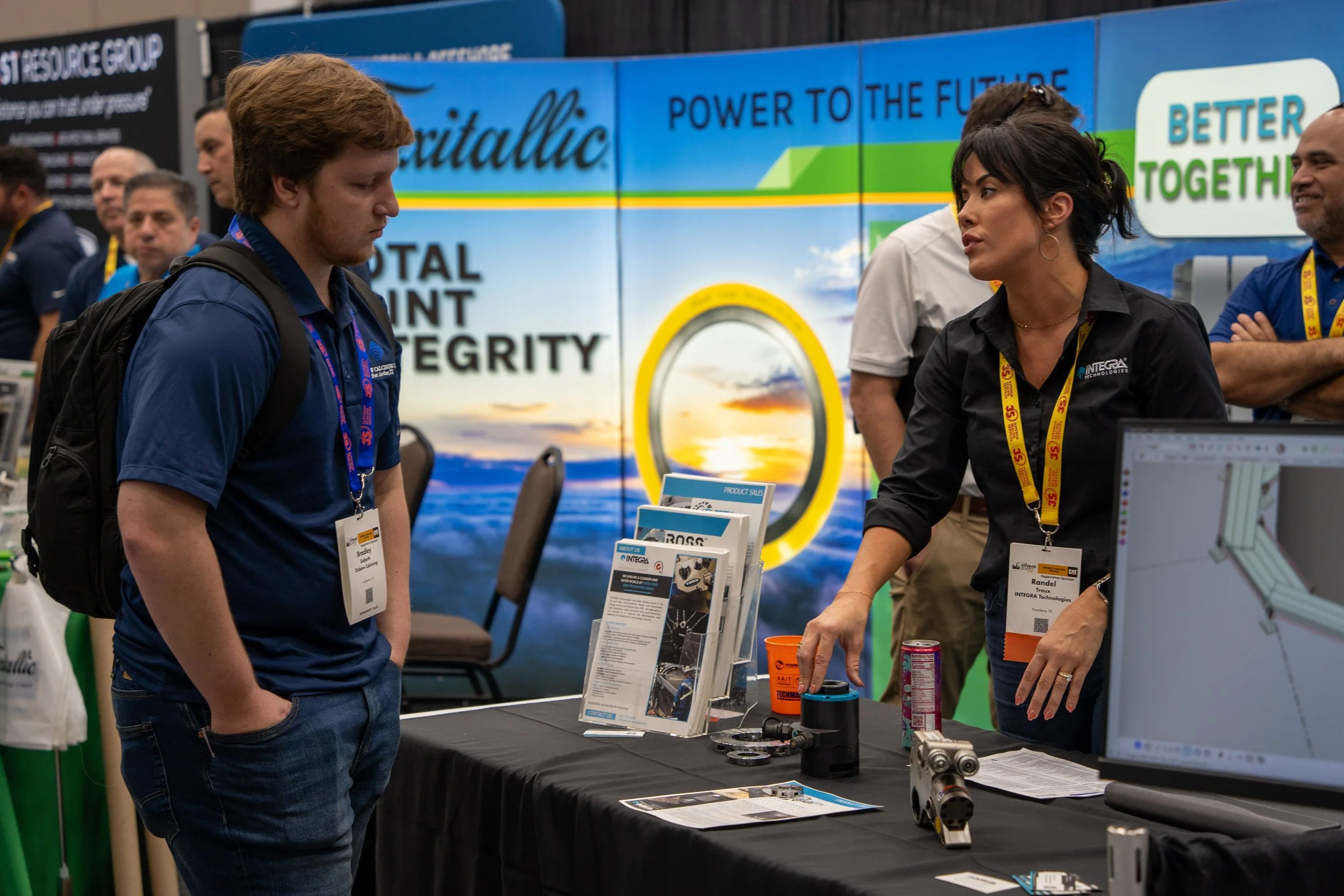 Two people talking at a trade show booth with promotional materials and machinery on display. The background features a large blue and green banner with text including 'Power to the Future' and 'Total Point Integrity'.