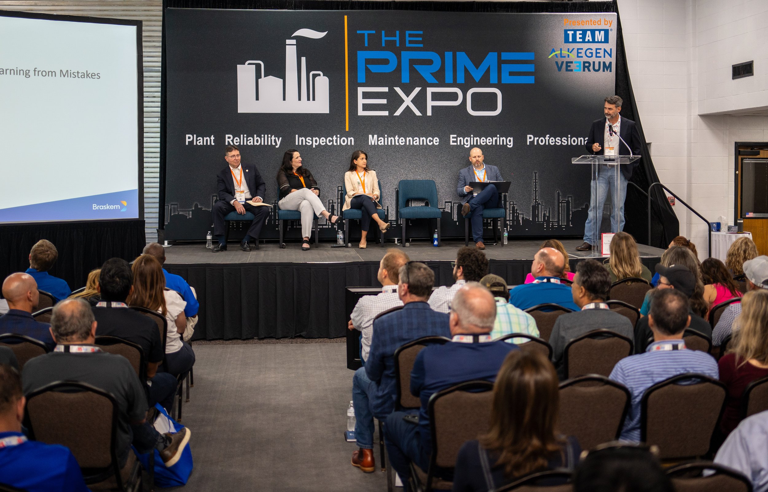 A panel of five people on stage at the Prime Expo, with four seated and one standing at a clear podium, addressing an audience. The backdrop features the event logo and topics such as plant reliability, inspection, maintenance, engineering, and profe