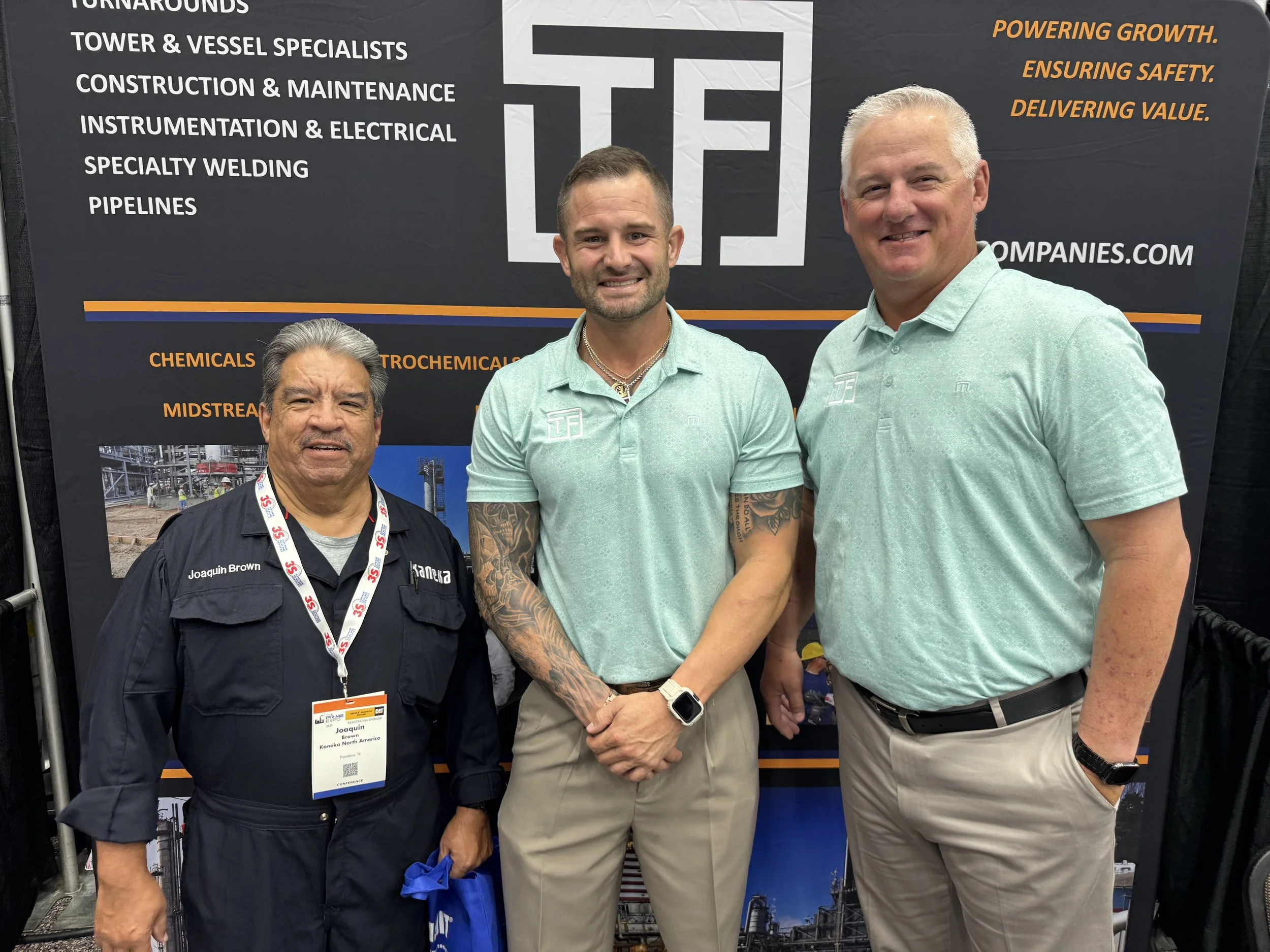 Three men standing in front of a black and orange industrial company banner at a trade show or conference. The banner lists services such as turnarounds, tower and vessel specialists, construction, maintenance, instrumentation, electrical, specialty 