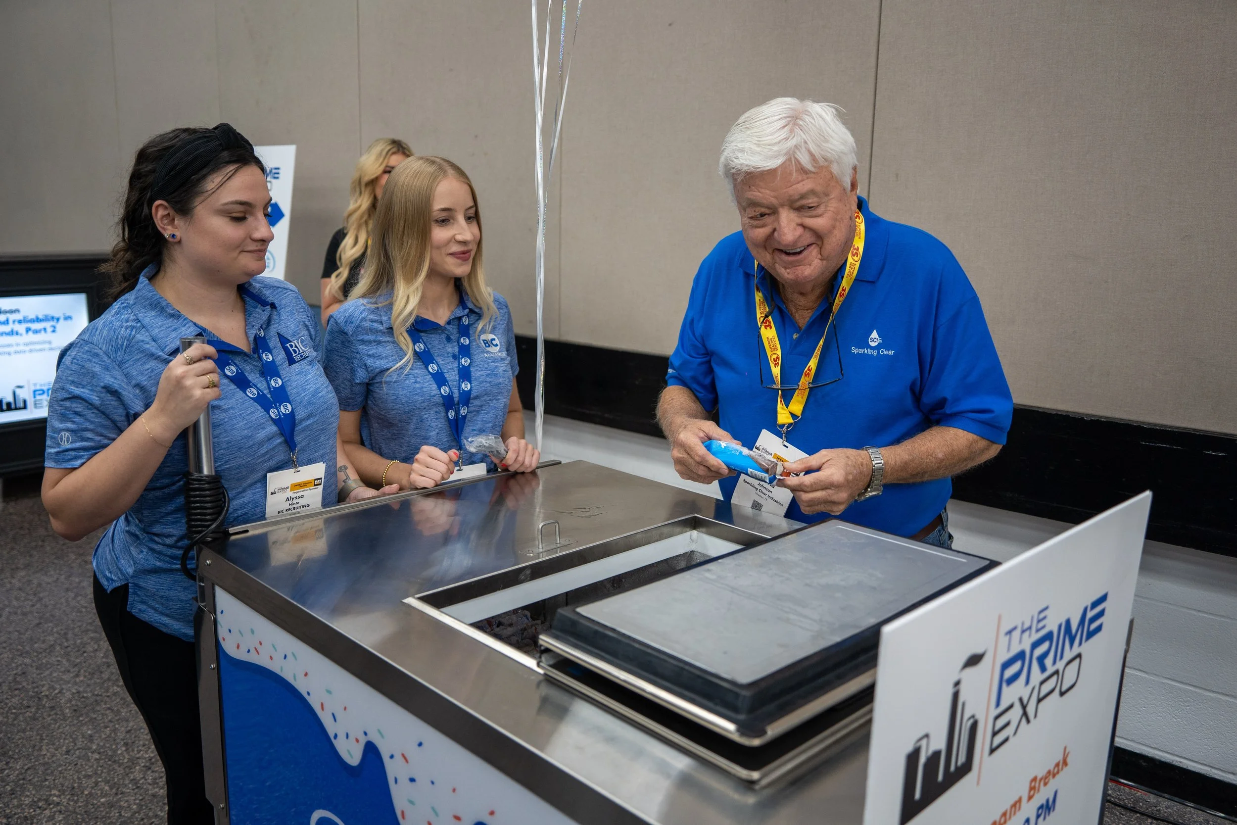 An older man in a blue polo shirt is smiling and holding a snack, standing in front of a silver cooler at an event called 'The Prime Expo' with three young women in blue shirts nearby. The women are watching and smiling.