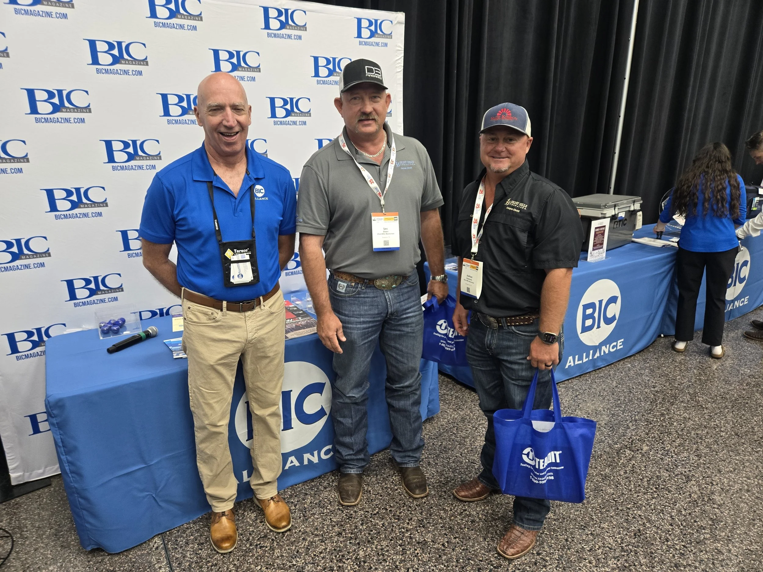 Three men standing at a booth with a BIC Alliance backdrop. Two are smiling at the camera, one looks serious. The man on the left is wearing a blue polo shirt and tan pants, the man in the middle is in a gray shirt and jeans, and the man on the right