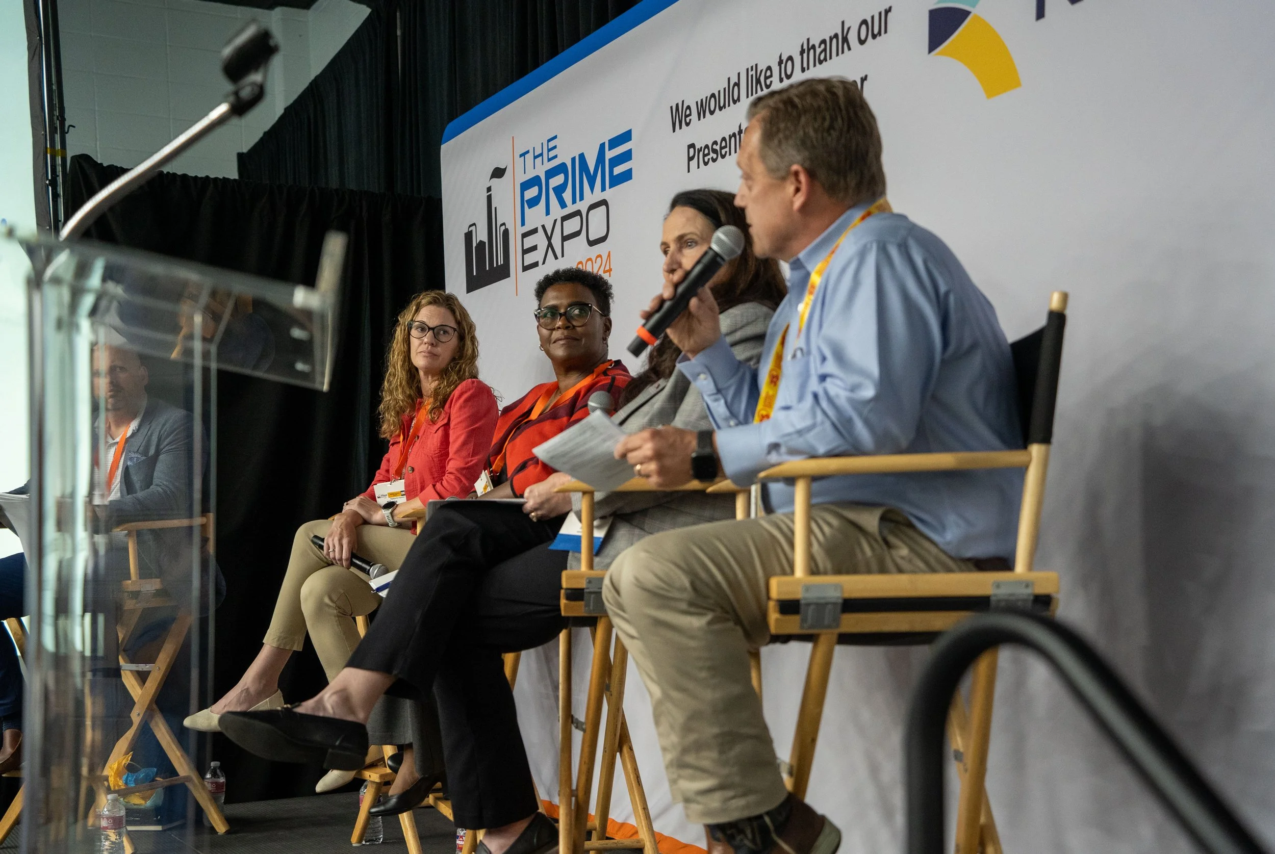 A panel of four people seated on a stage at The Prime Expo 2024, with a banner behind them displaying the event's logo and name. One person is speaking into a microphone while the others listen.