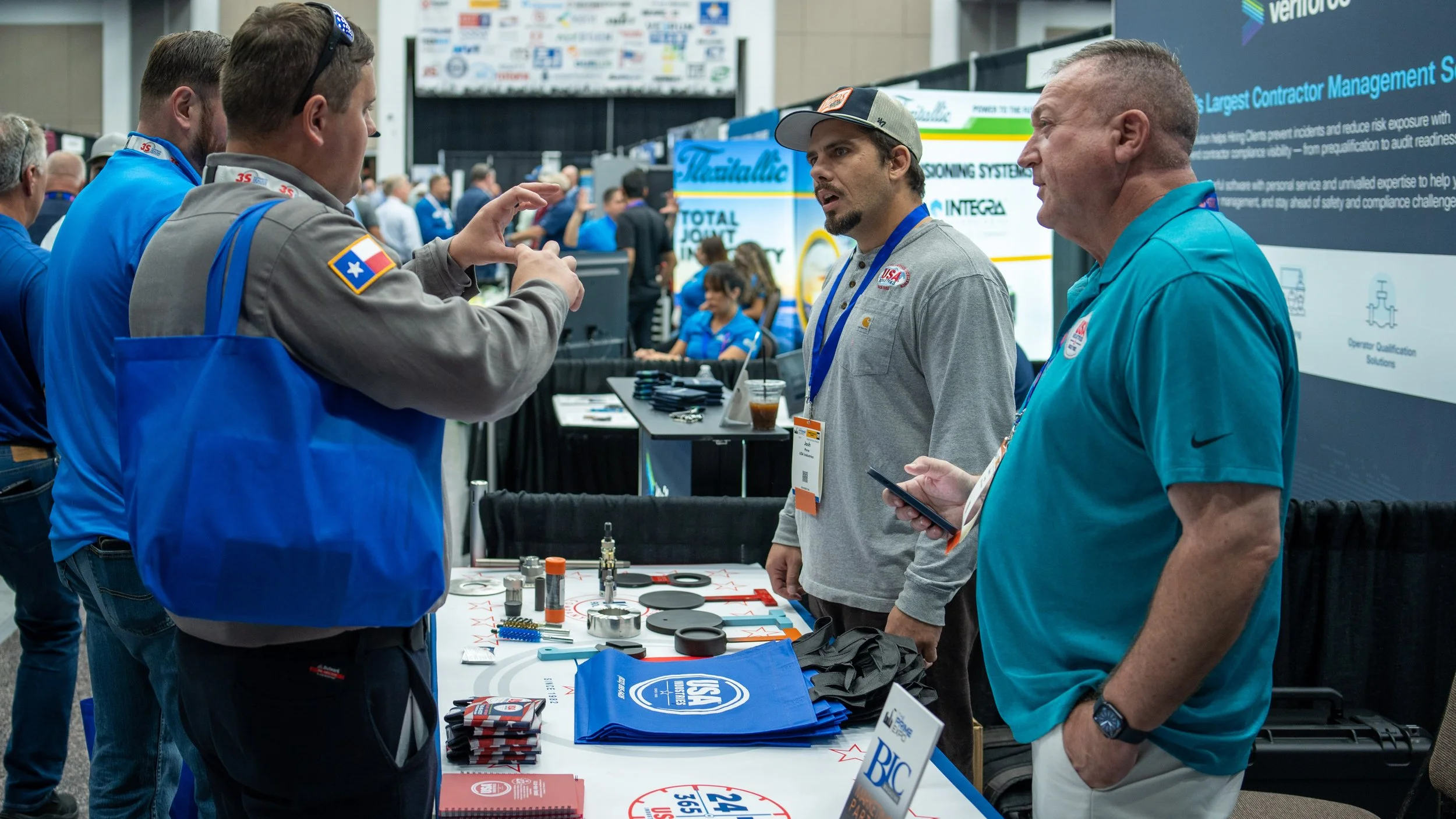 People at a trade show or conference booth discussing products, with various items and promotional materials on the table.