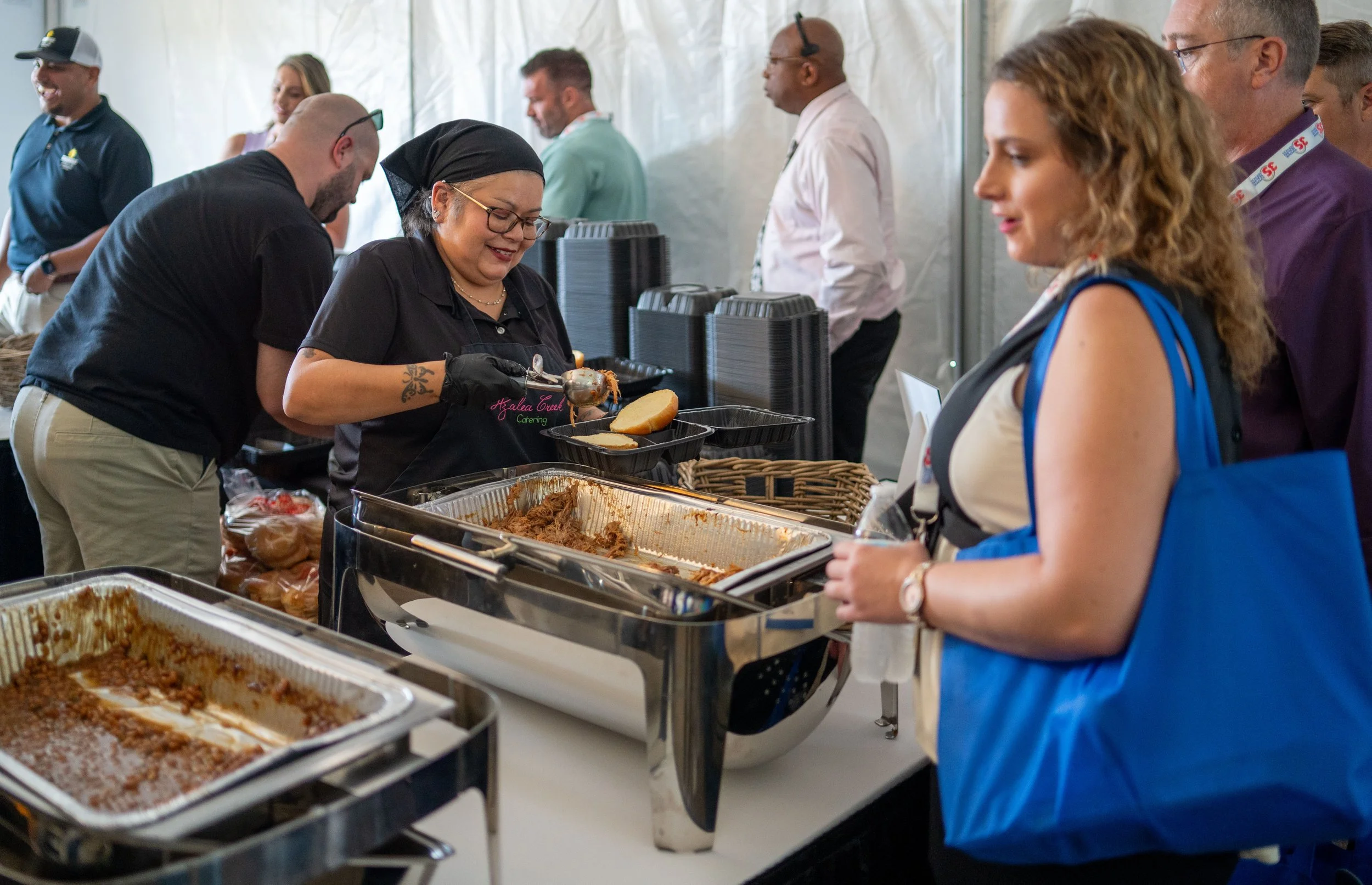 People in line at a buffet-style food service, with a woman serving food onto a plate from a chafing dish, while others wait in line and chat.