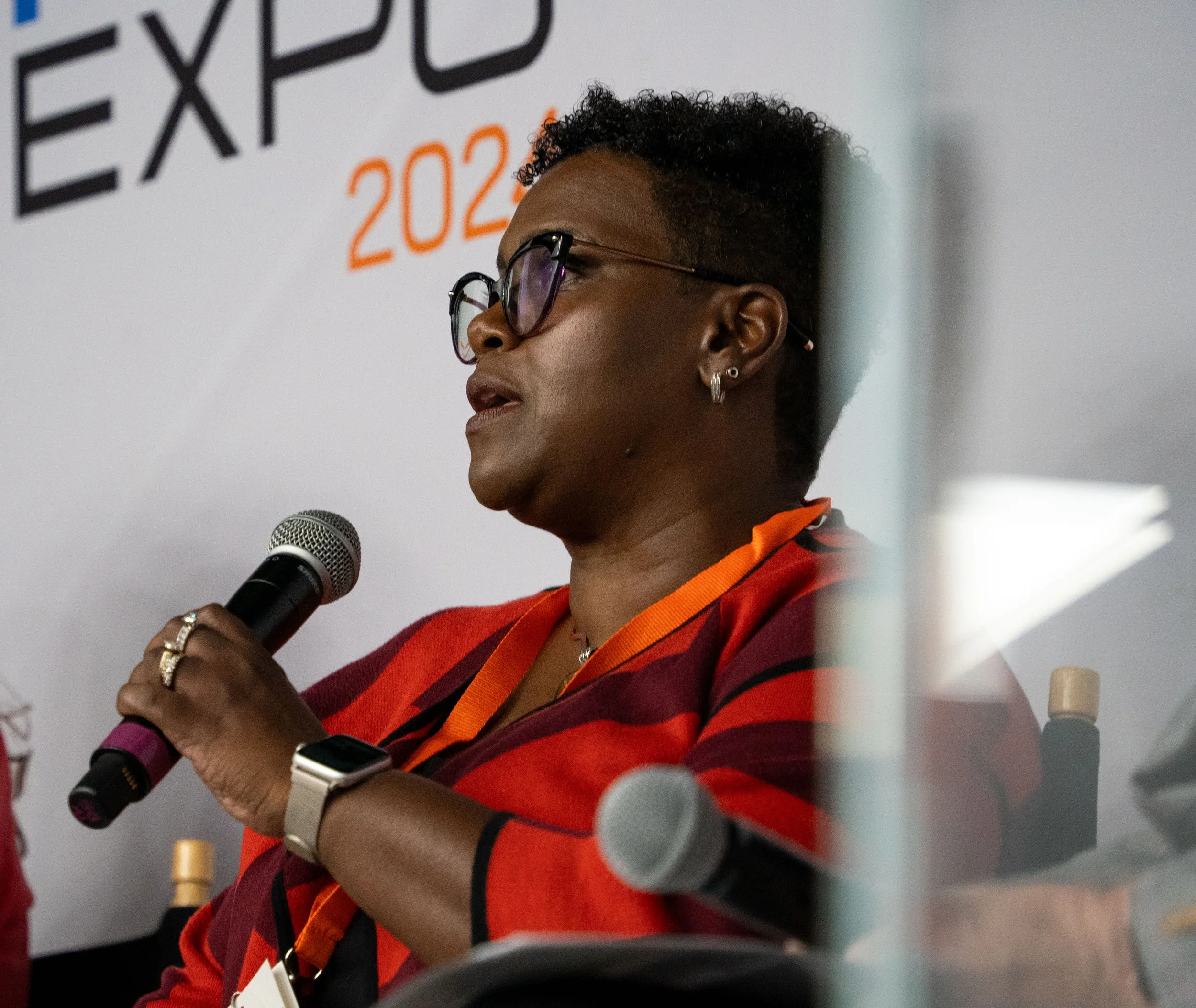 A woman speaking into a microphone during a conference or event. She is wearing glasses, earrings, a red and black striped blazer, and has a smartwatch on her left wrist. There is a banner behind her with the partial word 'EXPO' and the year 2023.