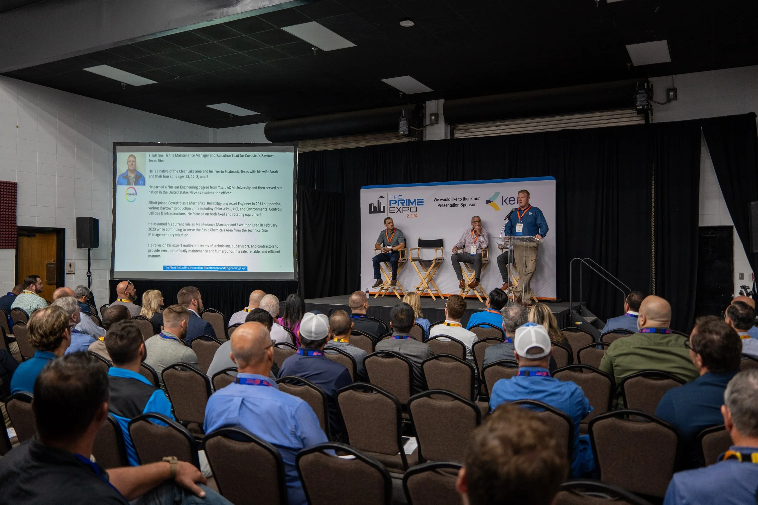 A conference room with an audience watching a stage presentation. The stage has three seated panelists and a speaker standing at a podium. A large screen displays a slide with a person's biography. Banners on the stage indicate the event is 'The Prim