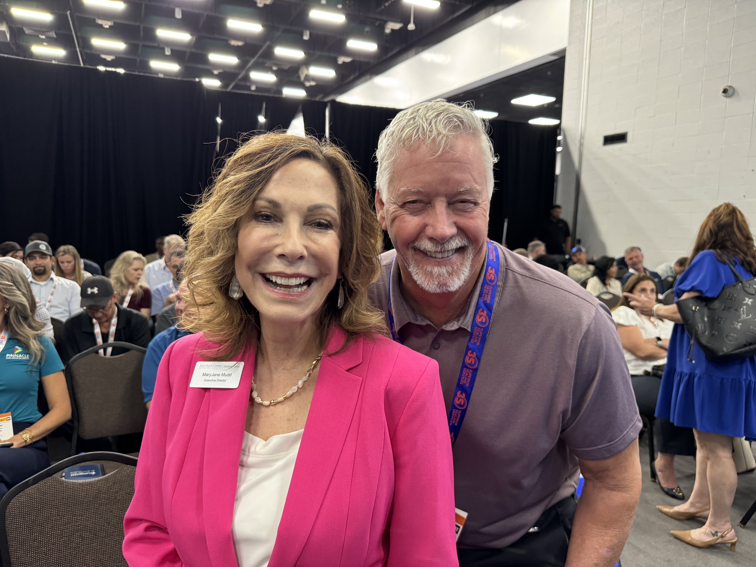 A woman in a pink blazer and a man in a lavender shirt smiling for a photo at a conference or event with many people seated and talking in the background.
