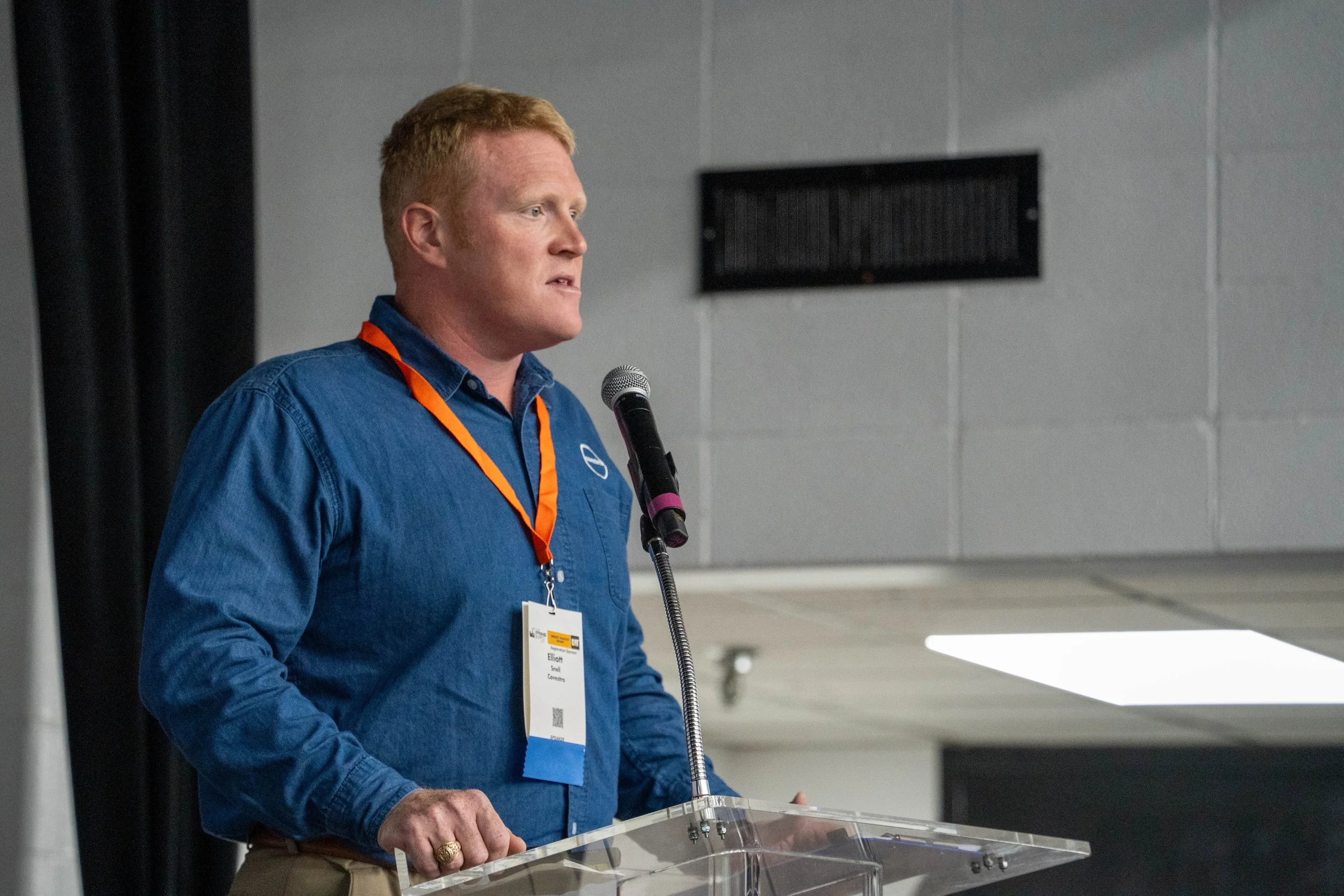A man with red hair and wearing a blue shirt standing at a clear podium, speaking into a microphone, in a conference room.