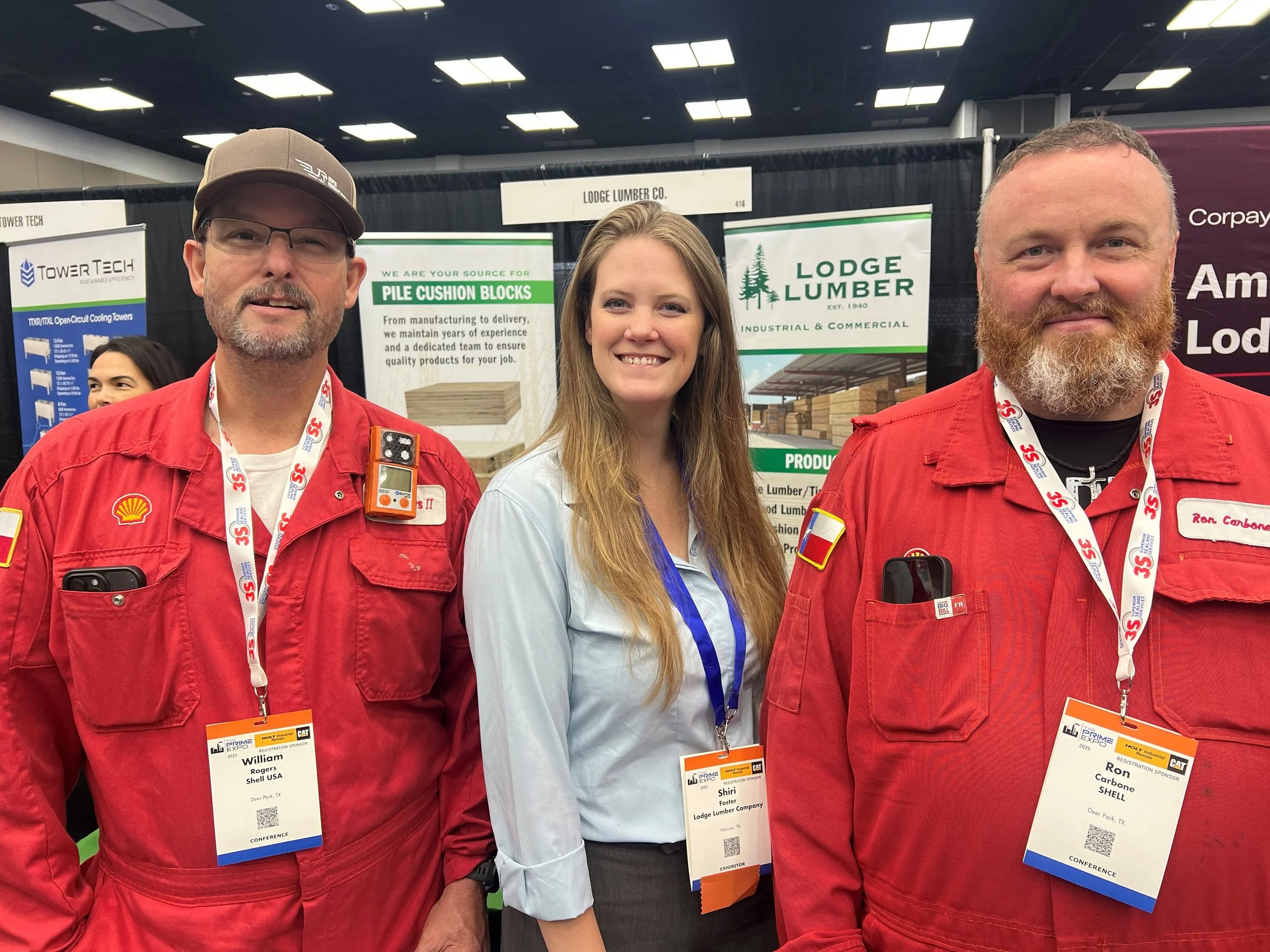 Three people standing together at a trade show, smiling at the camera. Two men wearing red work uniforms with name tags and badges, and a woman in a light blue shirt with a conference badge. Behind them are signs for lumber companies, and the setting