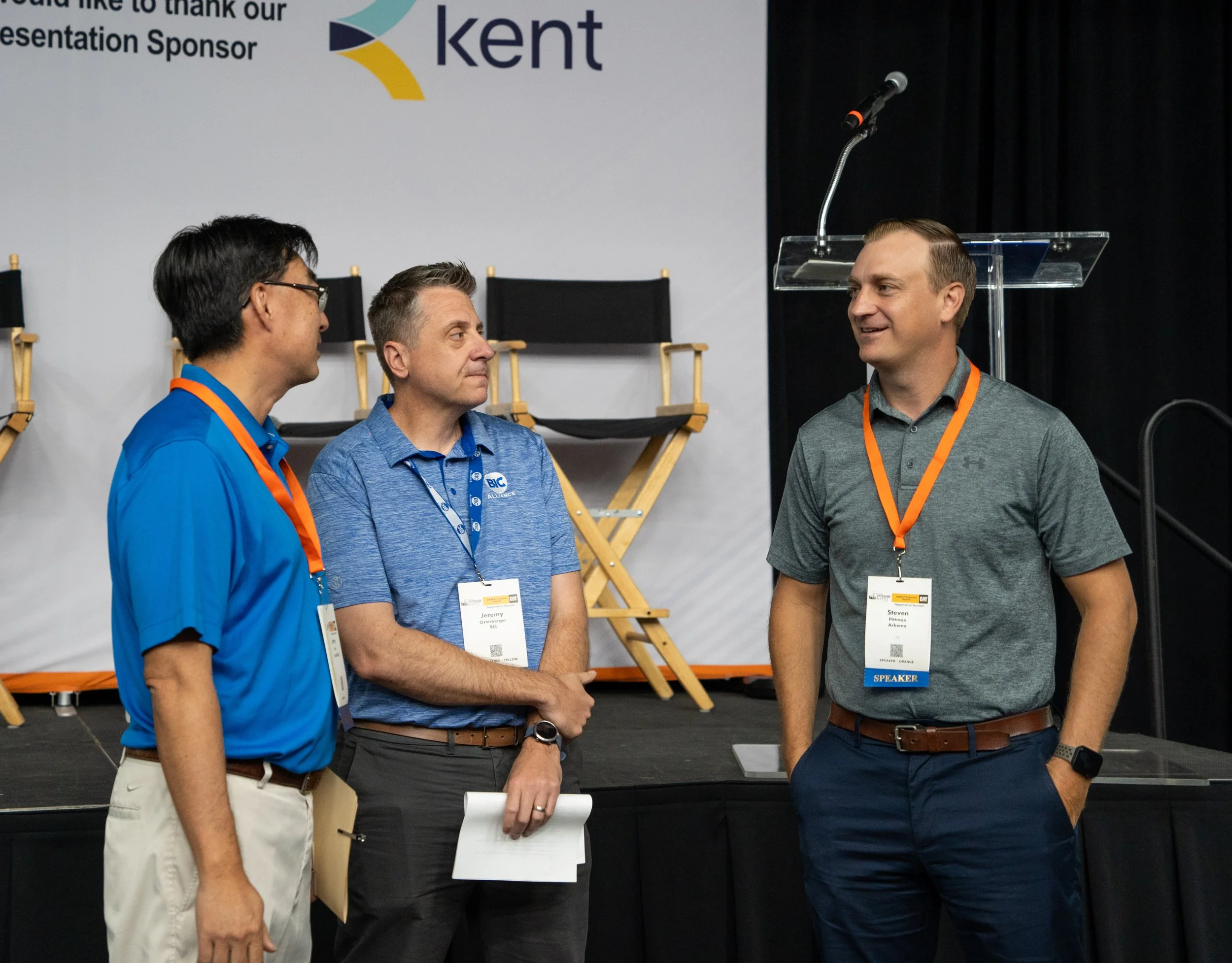Three men engaged in conversation at a conference, standing in front of a stage with a large banner displaying the words 'Kent' and a microphone on a stand. All three are wearing conference badges with orange lanyards.