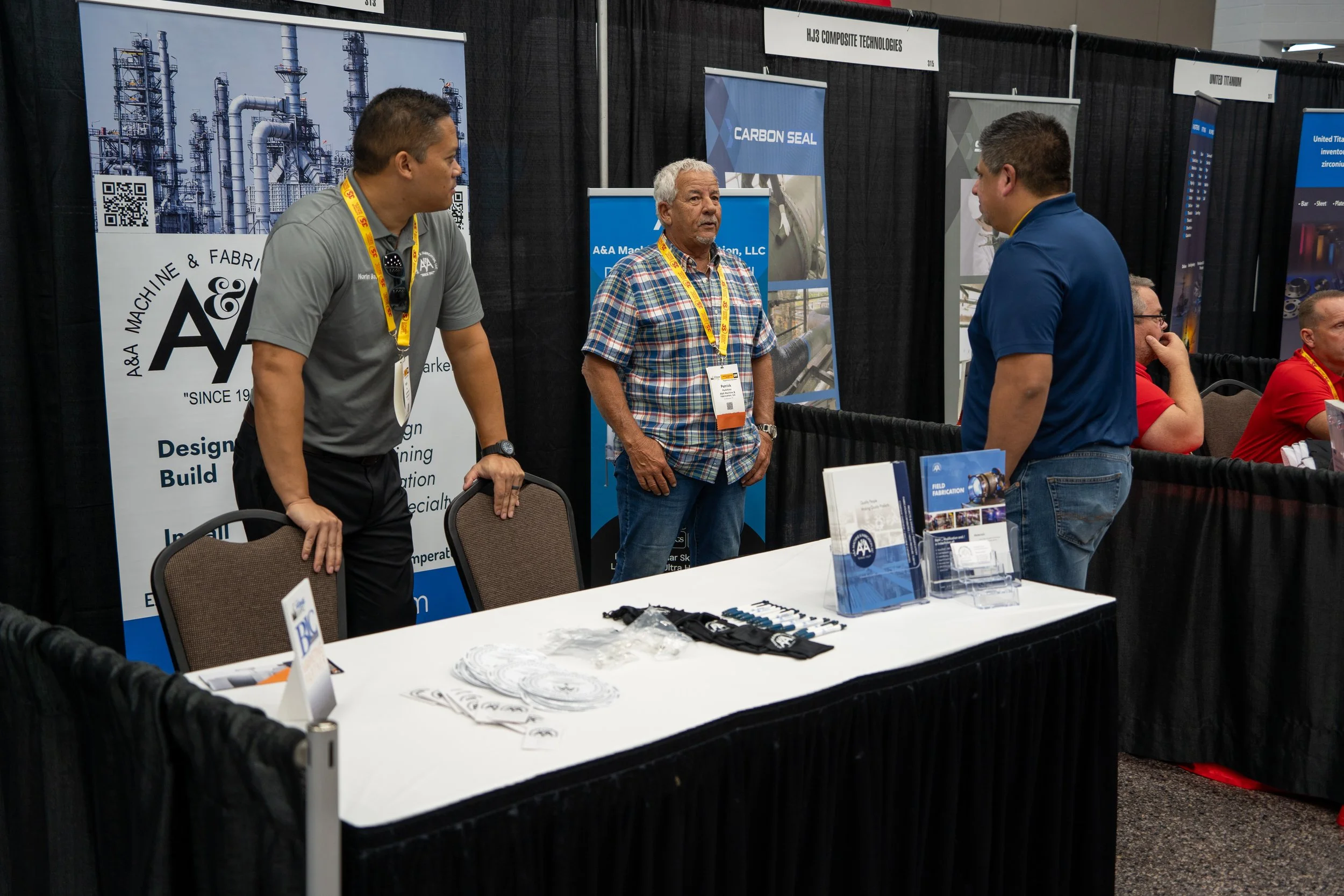 Three men are standing and talking at a booth during a convention or trade show. The booth has banners and informational materials displayed, with two tables holding brochures and promotional items. Two men sit at another table to the right, and a fe