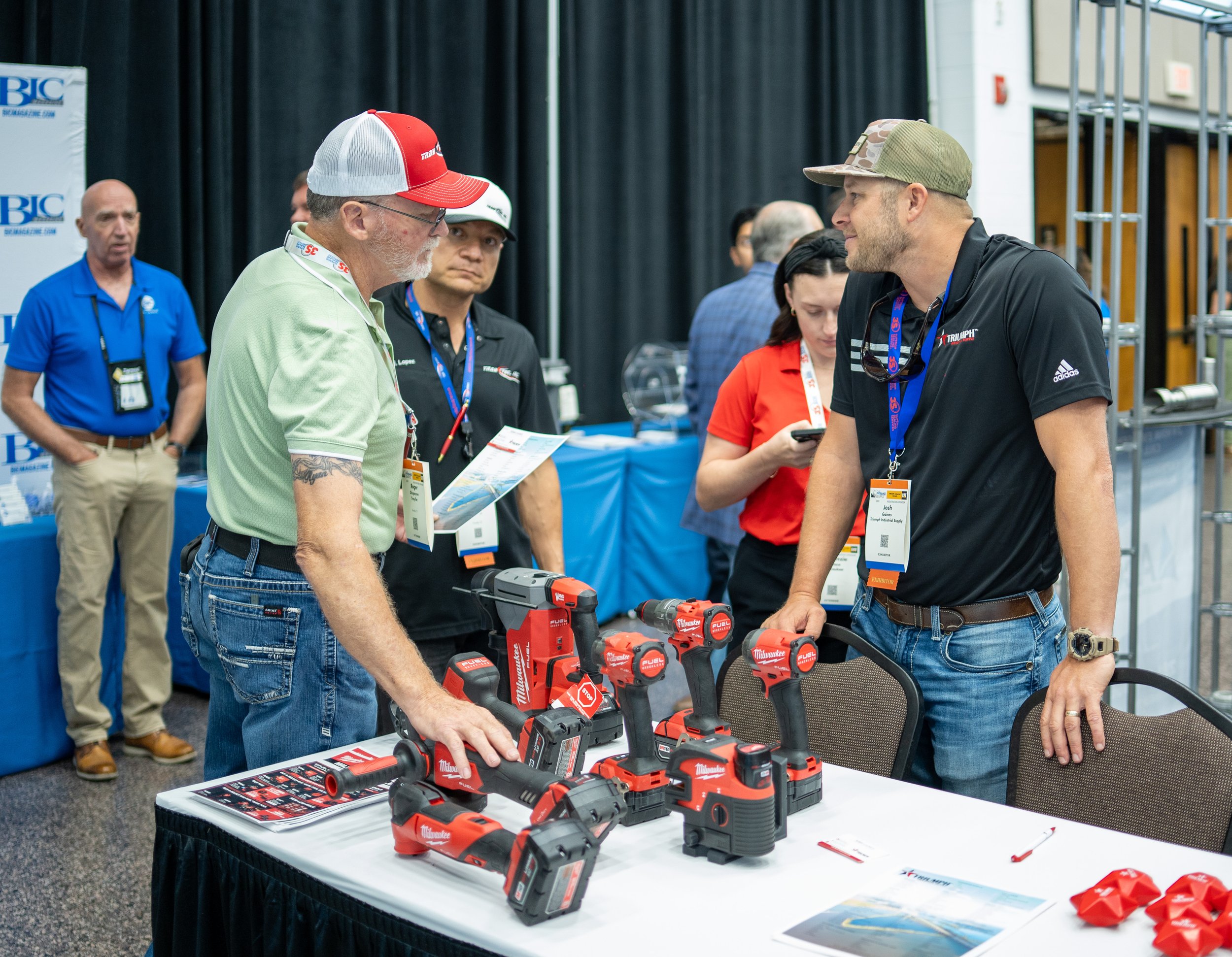Two men at a trade show booth discussing Milwaukee tools, with various power tools on display on the table.