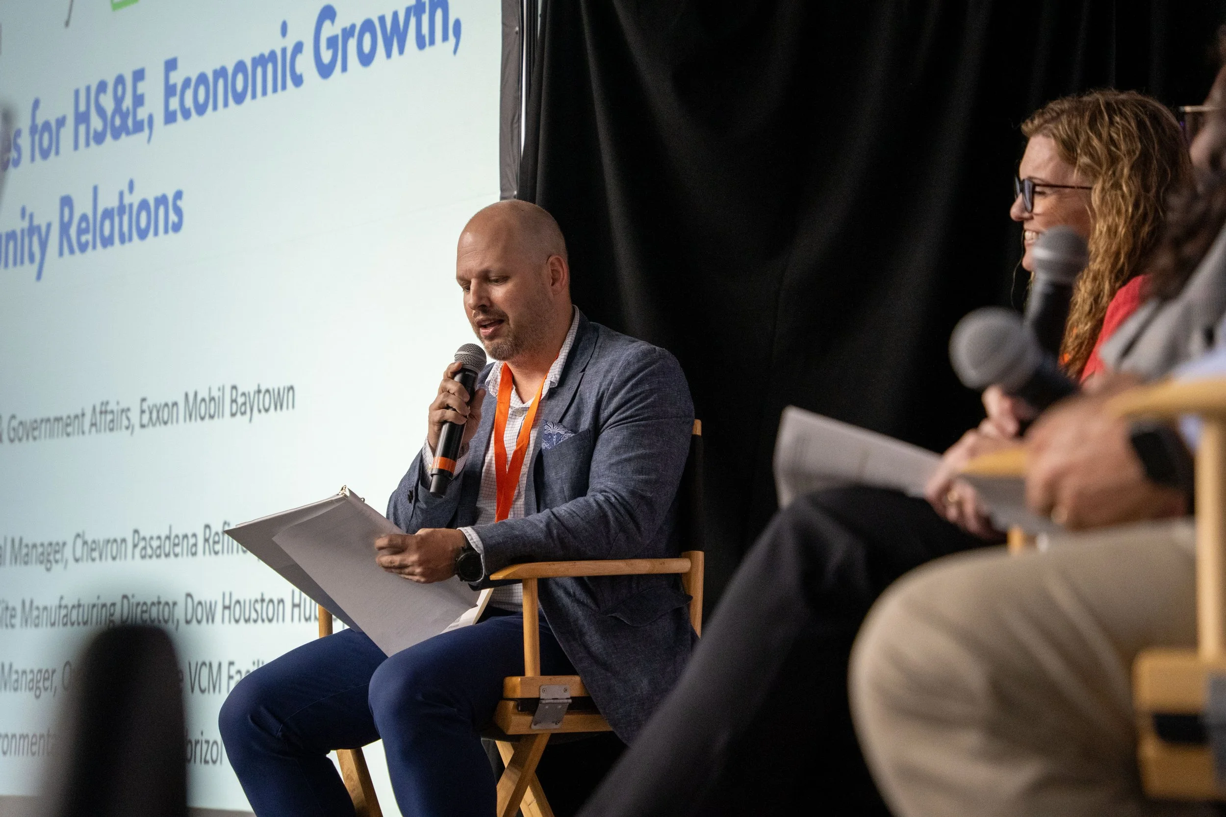 A man in a blue blazer speaking into a microphone while seated on a wooden chair during a panel discussion at a conference. Two women, also holding microphones and reading notes, are partially visible on the right. A large presentation slide with blu