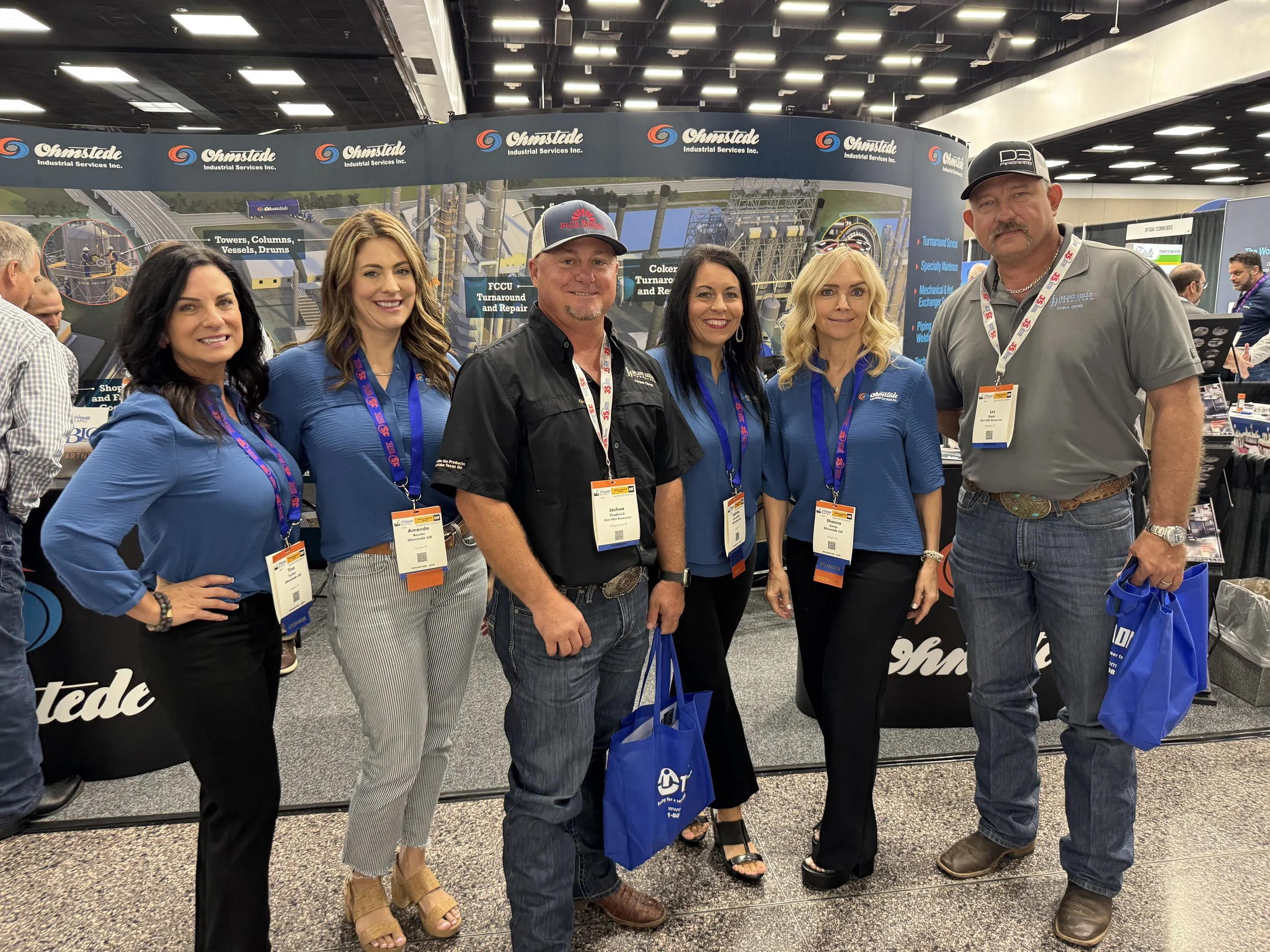 Group of six people standing together at a trade show booth, smiling for the camera. The booth features banners with the name Omstadle Industrial Services Inc. and machinery images in the background.