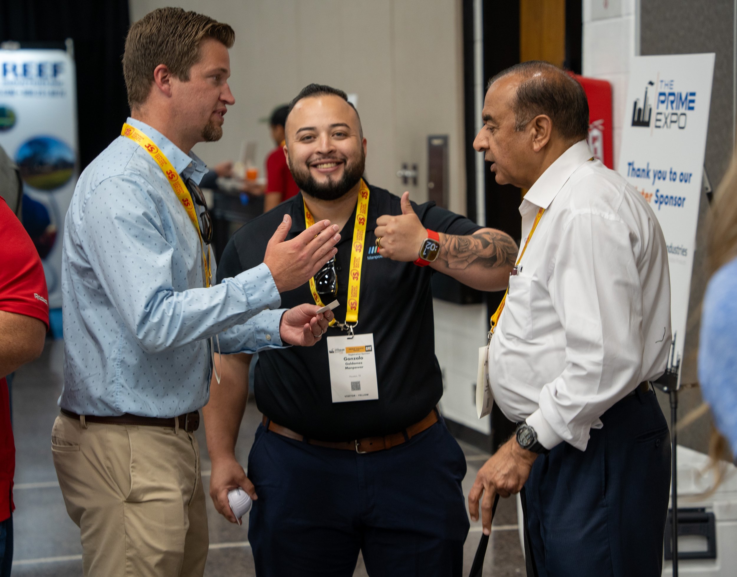 Three men are talking at a conference, wearing name badges and lanyards. The man on the right gestures while the man on the left listens, and the man in the middle smiles.