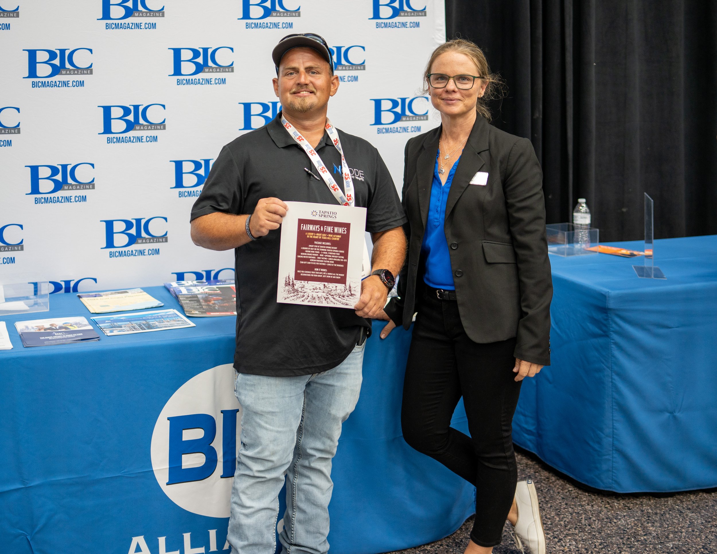 A man and a woman standing together at a promotional booth for BIC Magazine. The man is holding a brochure and wearing a black polo shirt and light jeans, while the woman is dressed in a black blazer, blue shirt, and black pants. They are smiling and