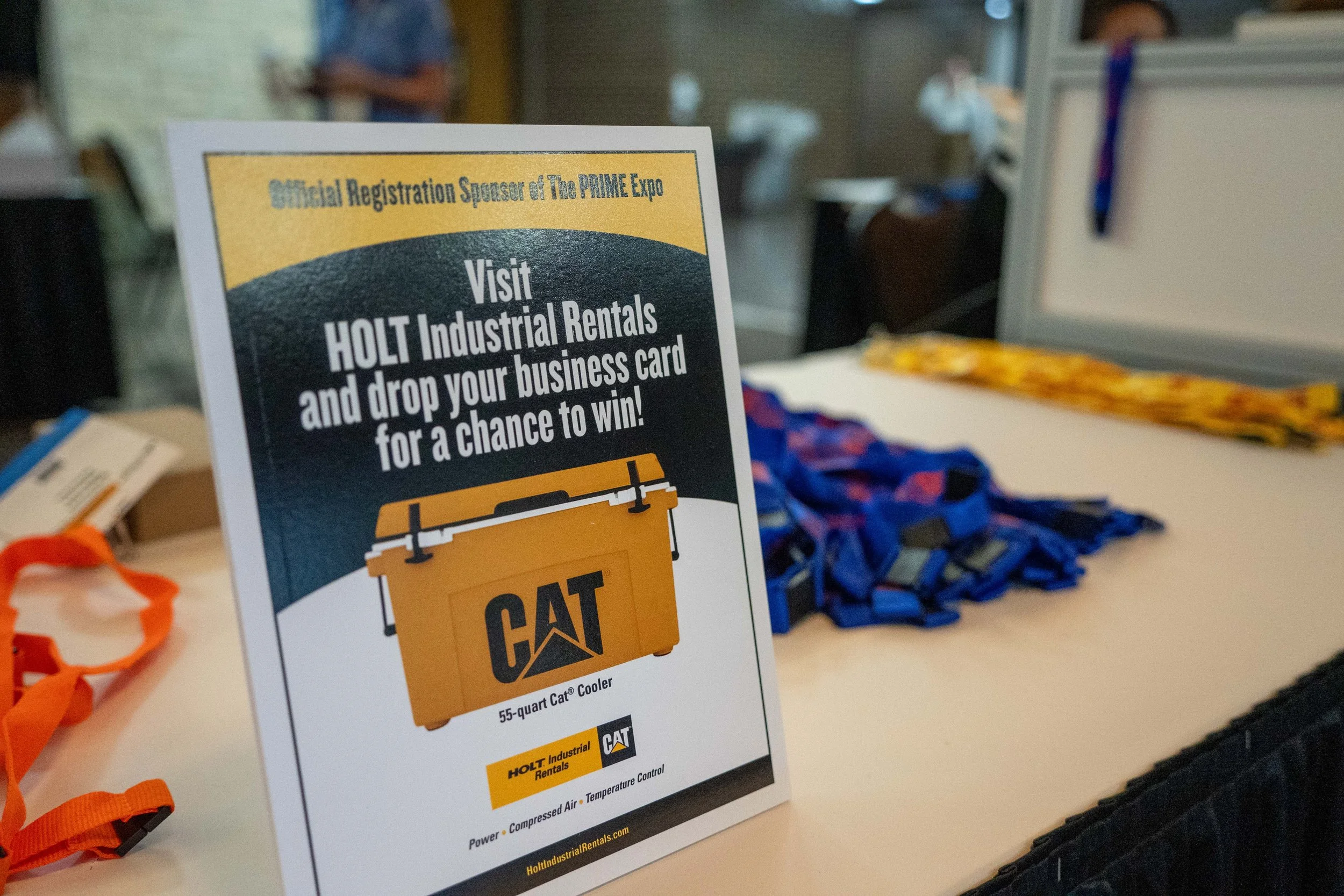 Sign at a booth advertising a chance to win a 55-quart CAT cooler by dropping a business card after visiting Holt Industrial Rentals, with raffle tickets and other items on the table.