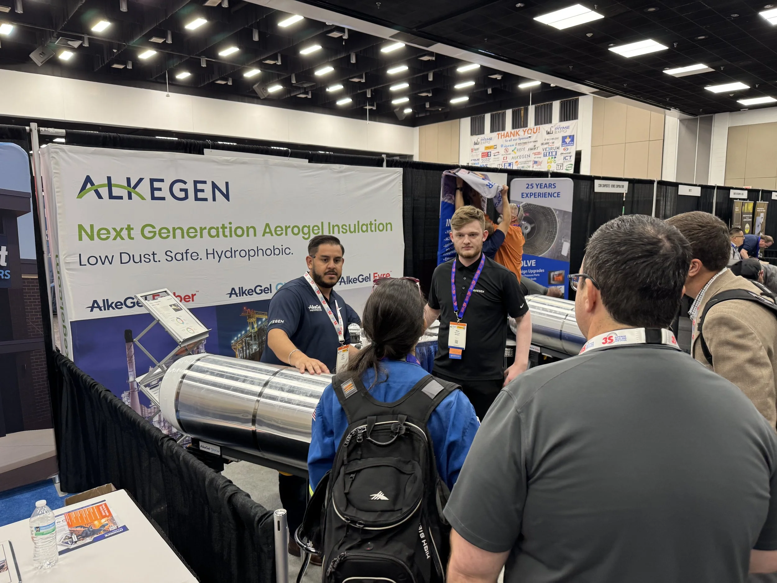 Group of people at a trade show booth for ALKEGEN, showcasing next-generation aerogel insulation. Two representatives stand behind the booth talking to visitors. The booth has a large banner with the ALKEGEN logo and information about low dust, safe,