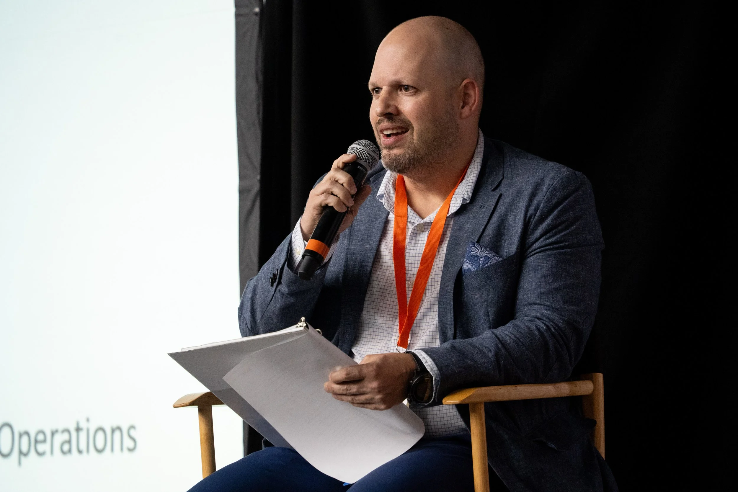 A bald man in a blue blazer and white shirt sitting on a chair, holding a microphone and some papers, speaking at an event with a presentation screen in the background.