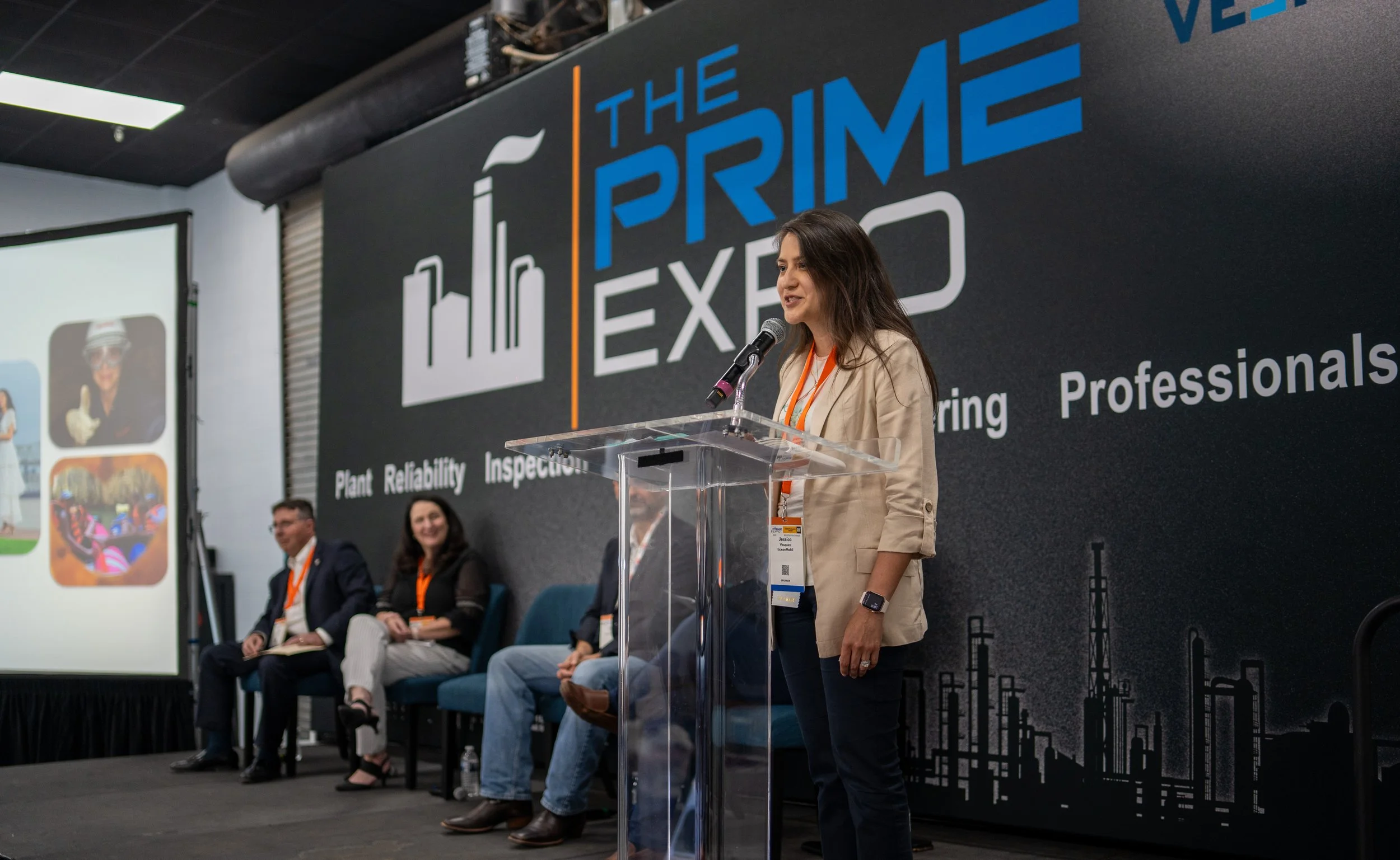 A woman speaking at a conference with a podium, with a large black banner behind her that reads "The Prime Expo" and lists words like Plant, Reliability, Inspection, and Professionals. There are three seated individuals on the stage, and a projection