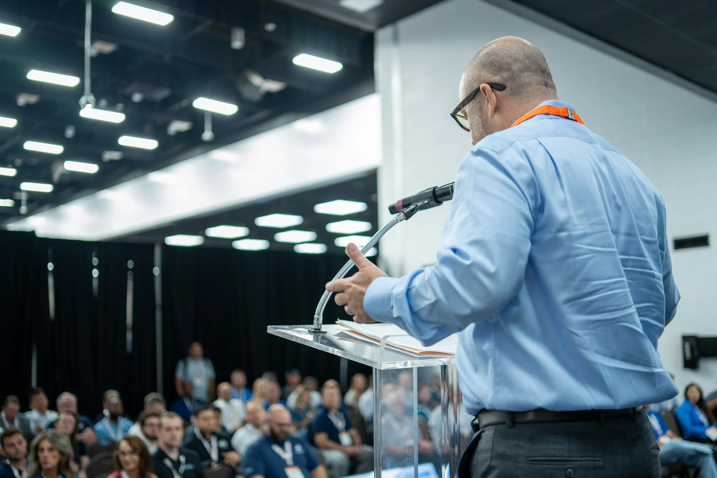 A man in a blue shirt and glasses giving a speech at a conference with an audience listening.