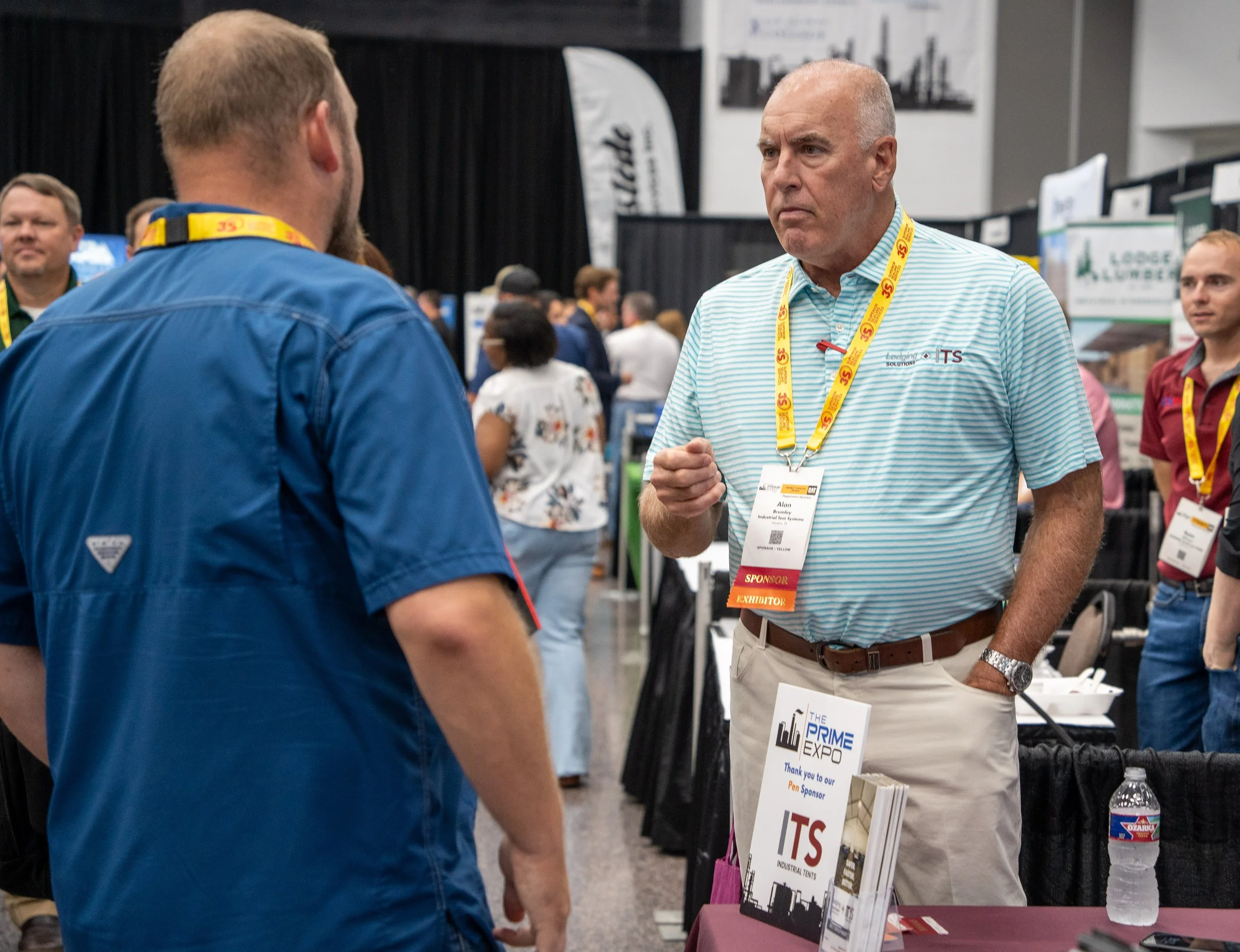 A man with a bald head wearing a blue and white striped polo shirt and glasses around his neck is speaking to another man with a beard and a blue shirt at a trade show. The man with the striped shirt has an exhibitor badge hanging around his neck, an