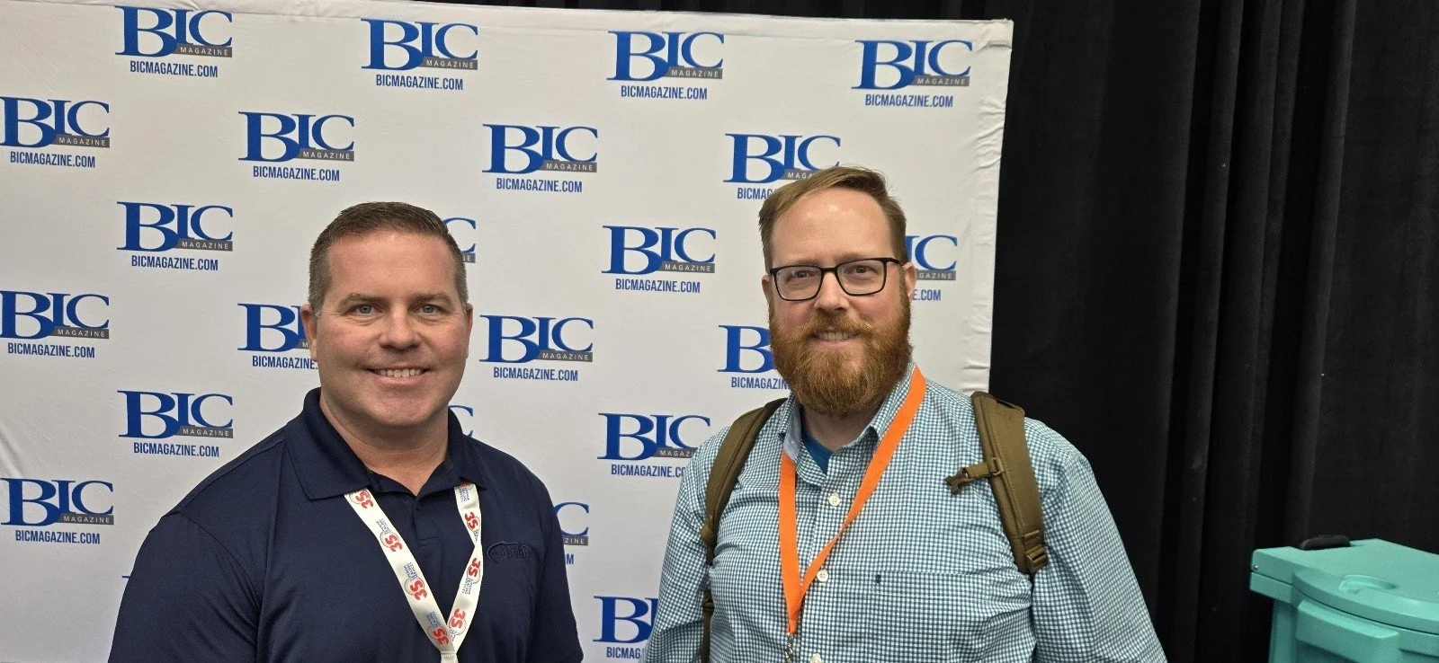 Two men smiling standing in front of a backdrop with the logo and website of BIC Magazine, one wearing a dark polo shirt and the other a checked shirt with a backpack.