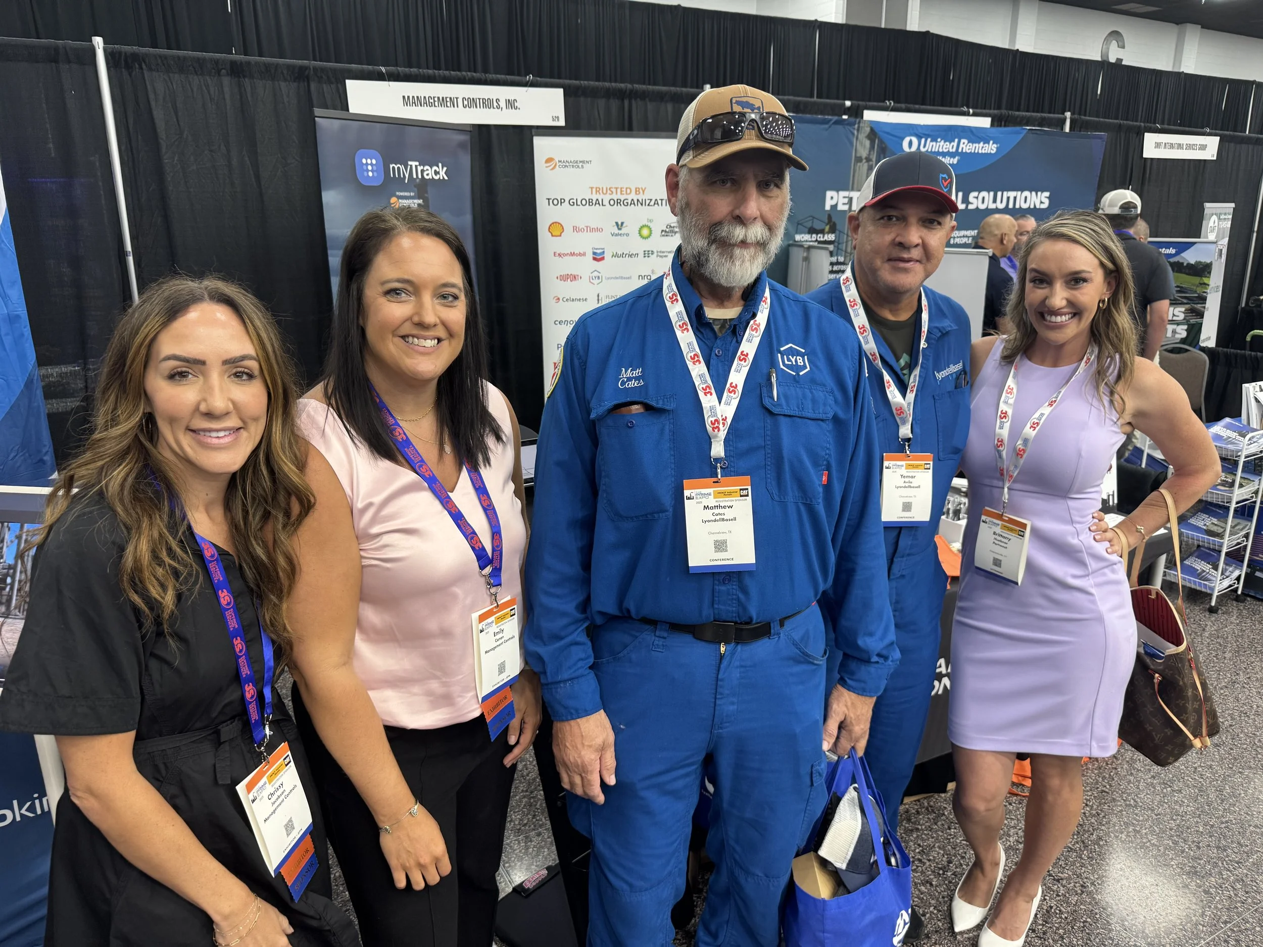 Group of five people, four women and one man, at a trade event or conference, wearing name tags and standing in front of exhibitor booths, smiling at the camera.