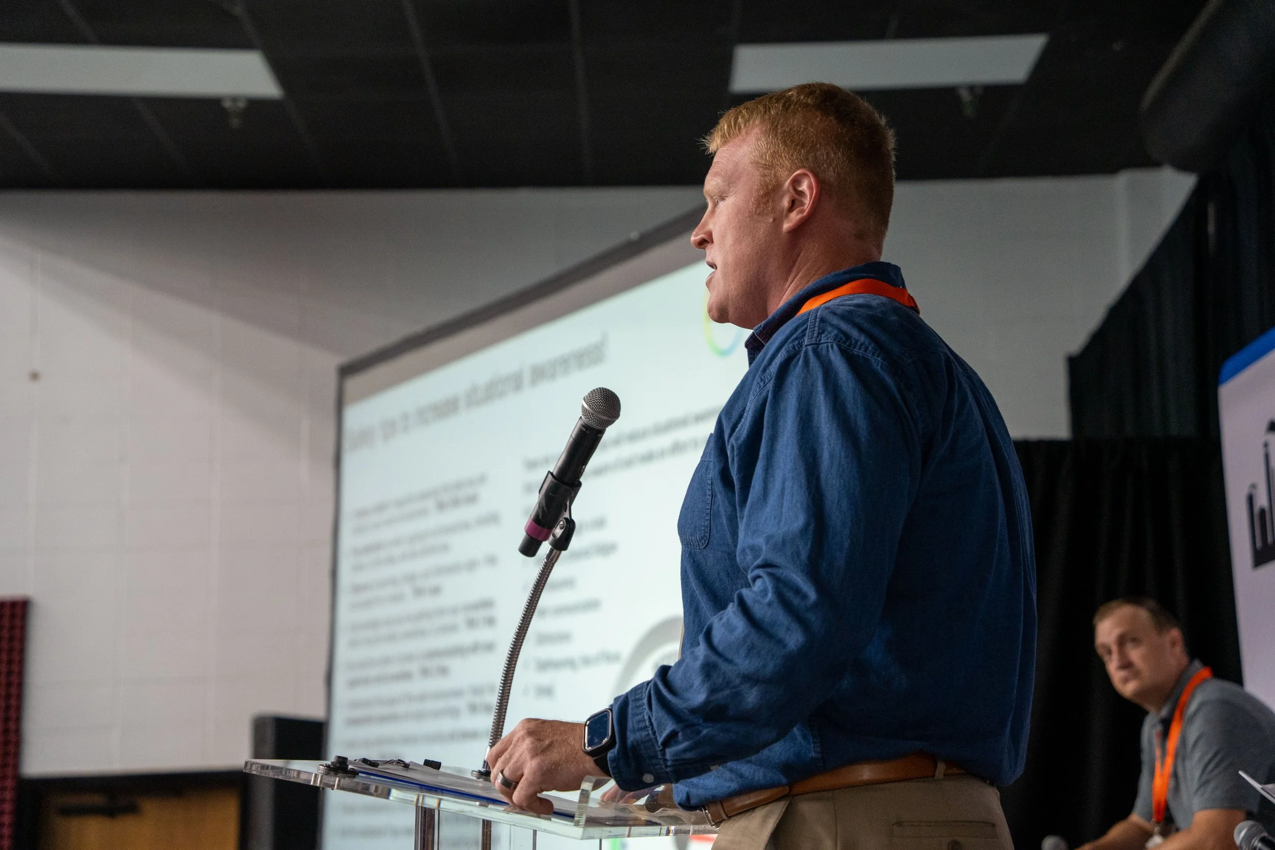 A man with red hair and a navy blue shirt speaking at a podium with a microphone during a presentation in a conference room. Another man is seated in the background, and a presentation slide is projected on the screen behind them.