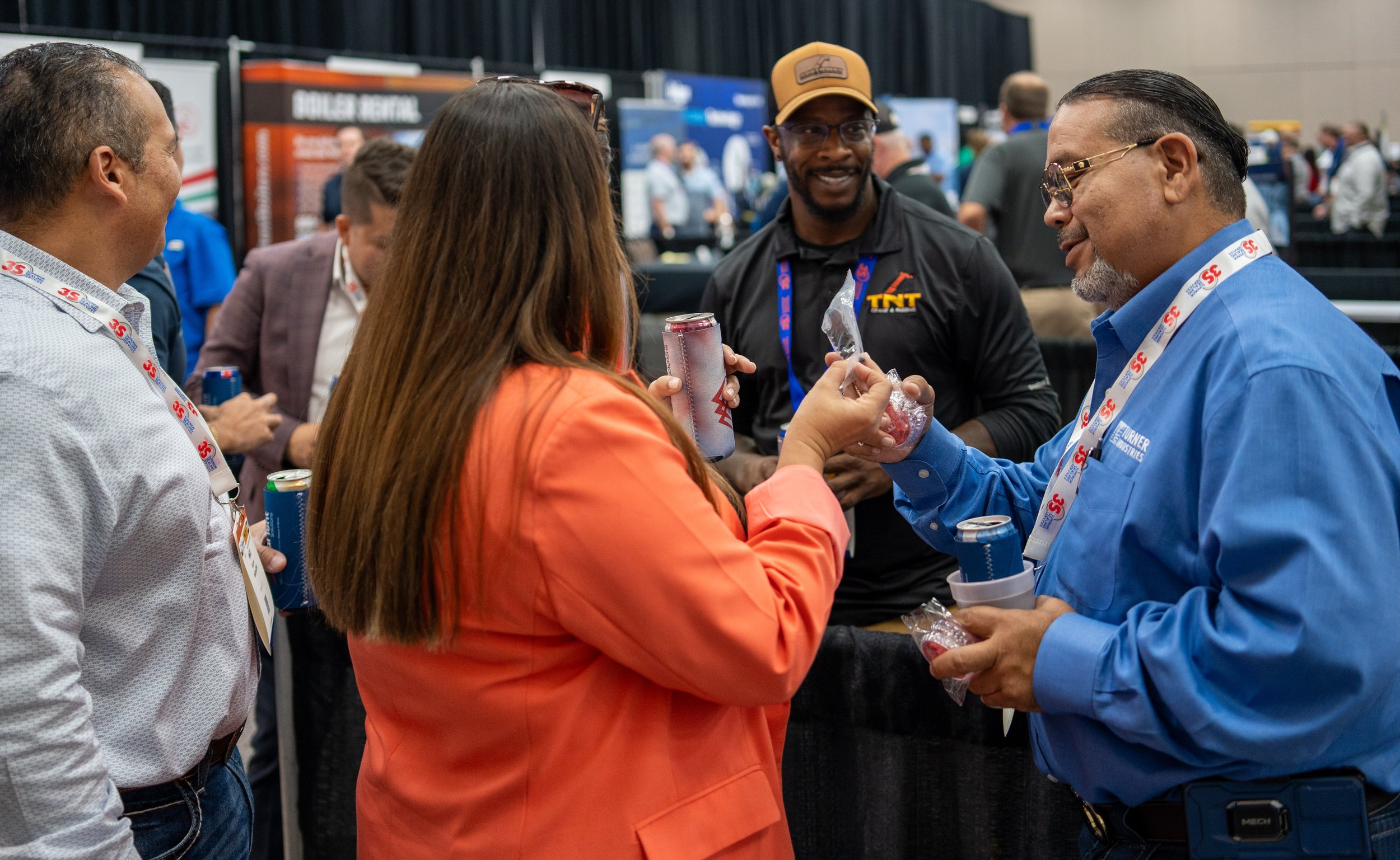Group of people chatting at a trade show or conference, holding beverages and promotional items, with booths and banners in the background.