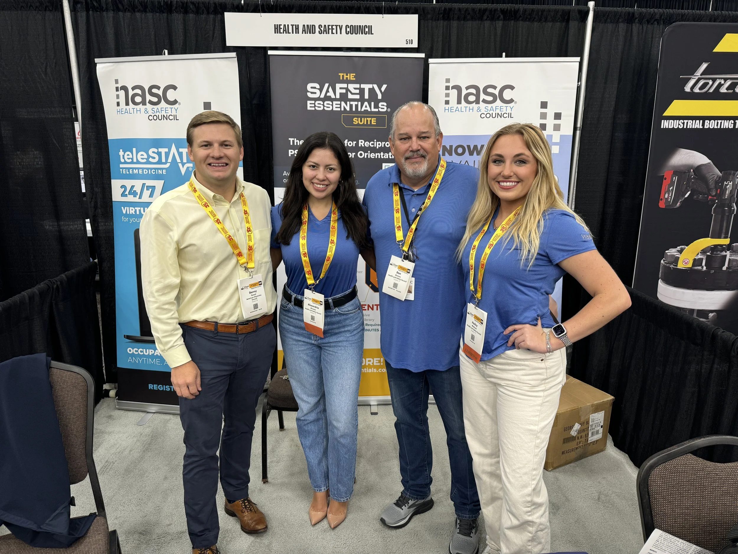 Four people standing together at a trade show booth, smiling for the photo, with banners behind them displaying 'HASC' and 'The Safety Essentials Suite,' and a sign that says 'Health and Safety Council' above.