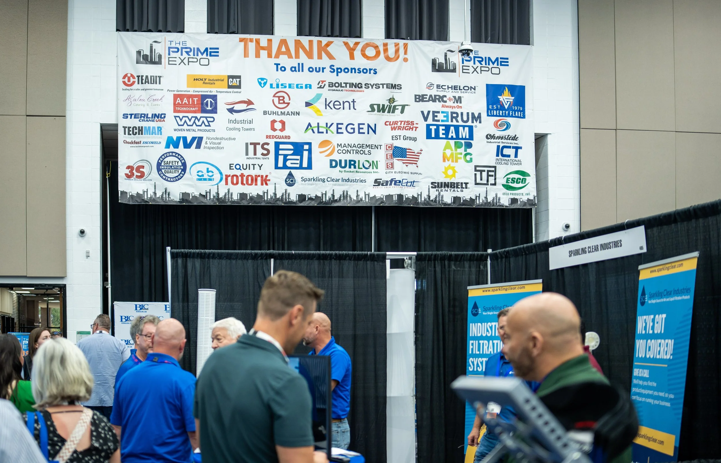 A large banner with sponsor logos and a 'Thank You' message at the Prime Expo, with people attending and engaging at booths below.
