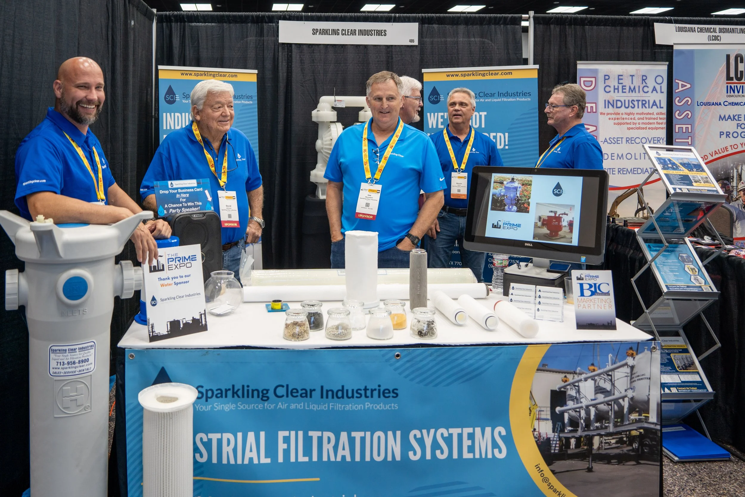 A group of five men wearing blue shirts with yellow lanyards standing behind a booth at a trade show, showcasing filtration systems. The booth has banners for Sparkling Clear Industries and displays various filters, pipes, and a monitor showing indus