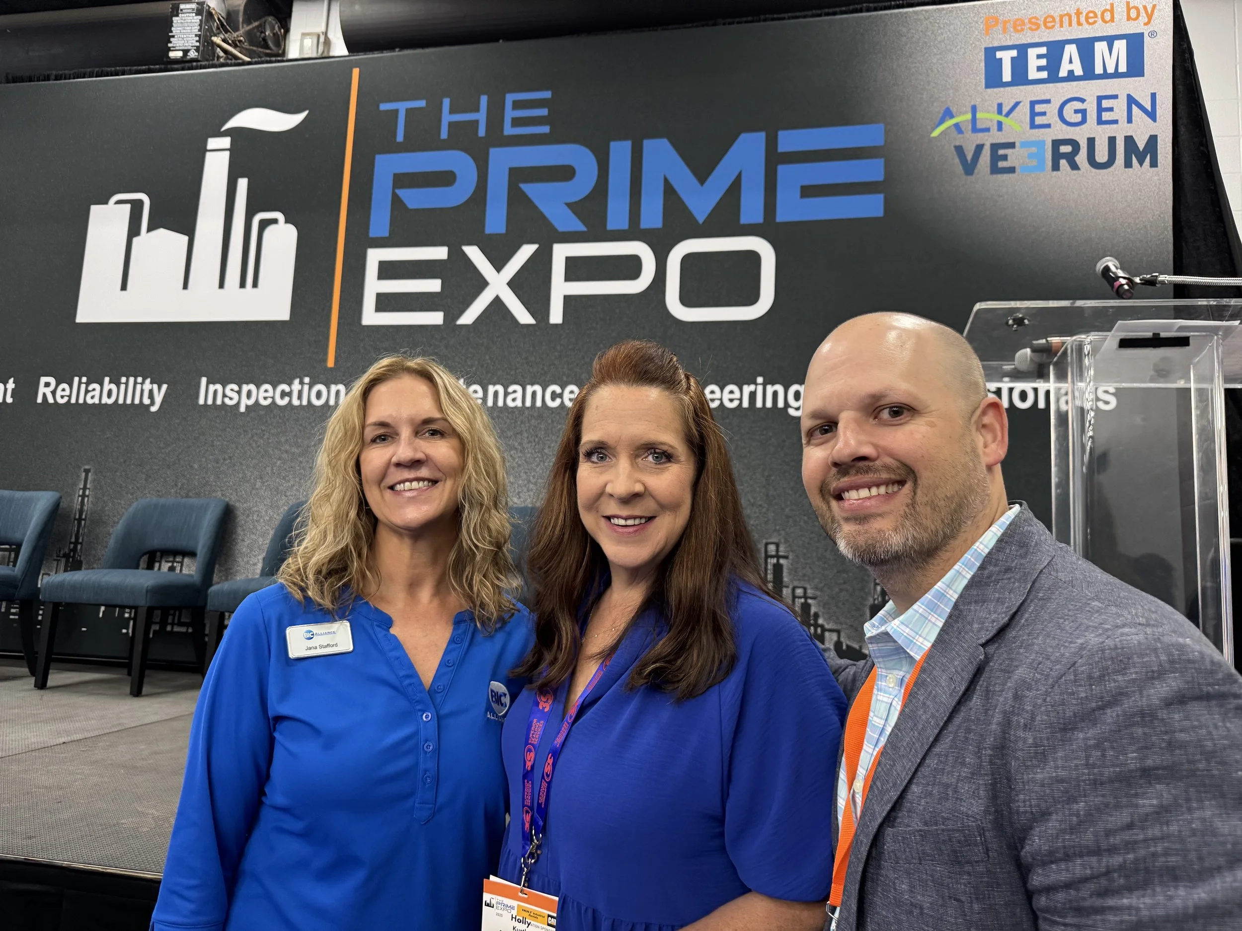 Three people smiling for a selfie at The Prime Expo event, with a backdrop displaying event information and logos.