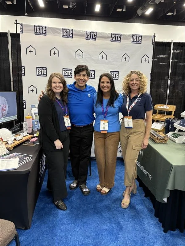 Four people standing together at an expo booth, smiling. They are wearing business casual attire with name tags. The booth has various informational materials and displays, with a backdrop featuring logos of ISTC and some icons.