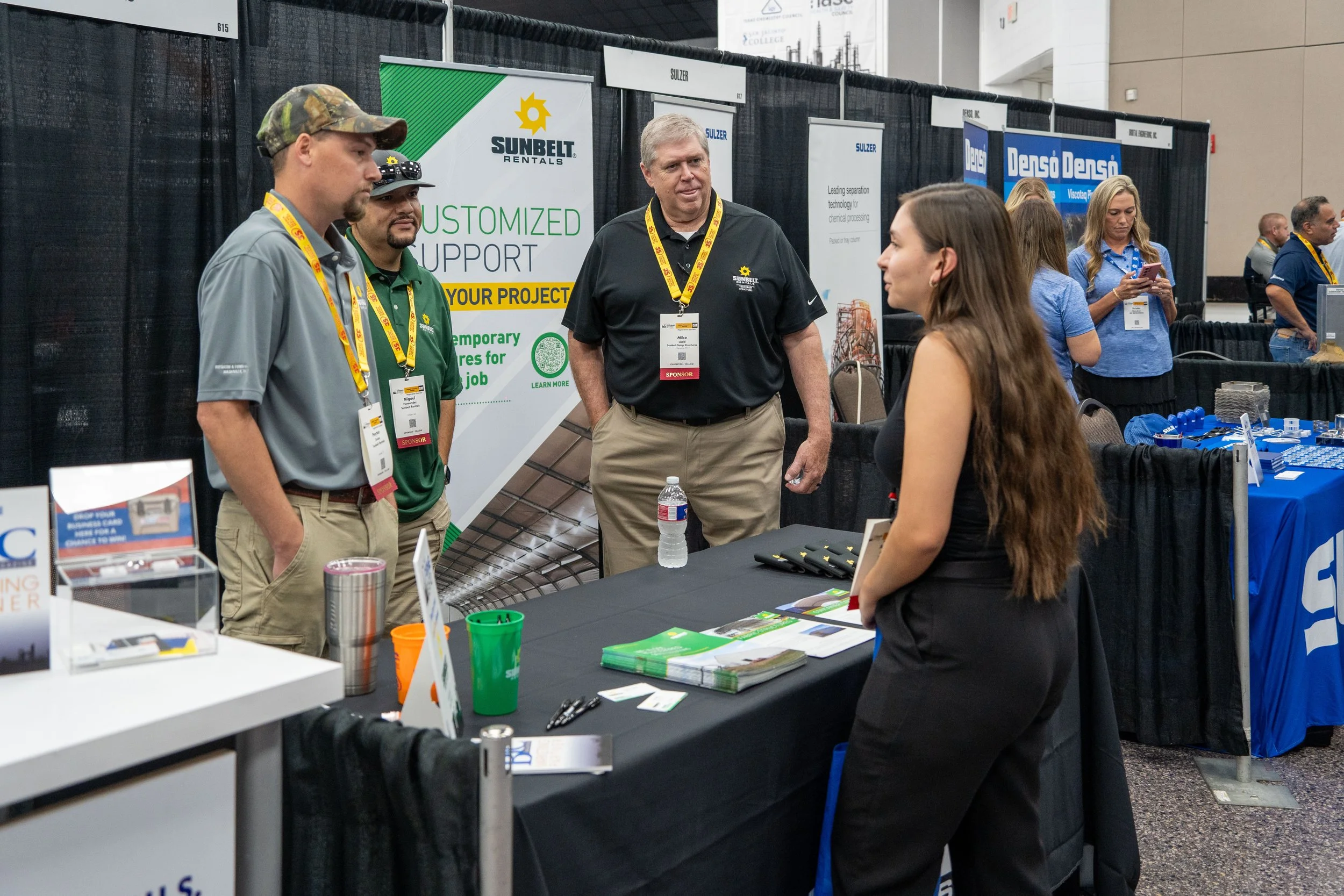 A woman is speaking with three men at a trade show booth, with various promotional materials and banners in the background.