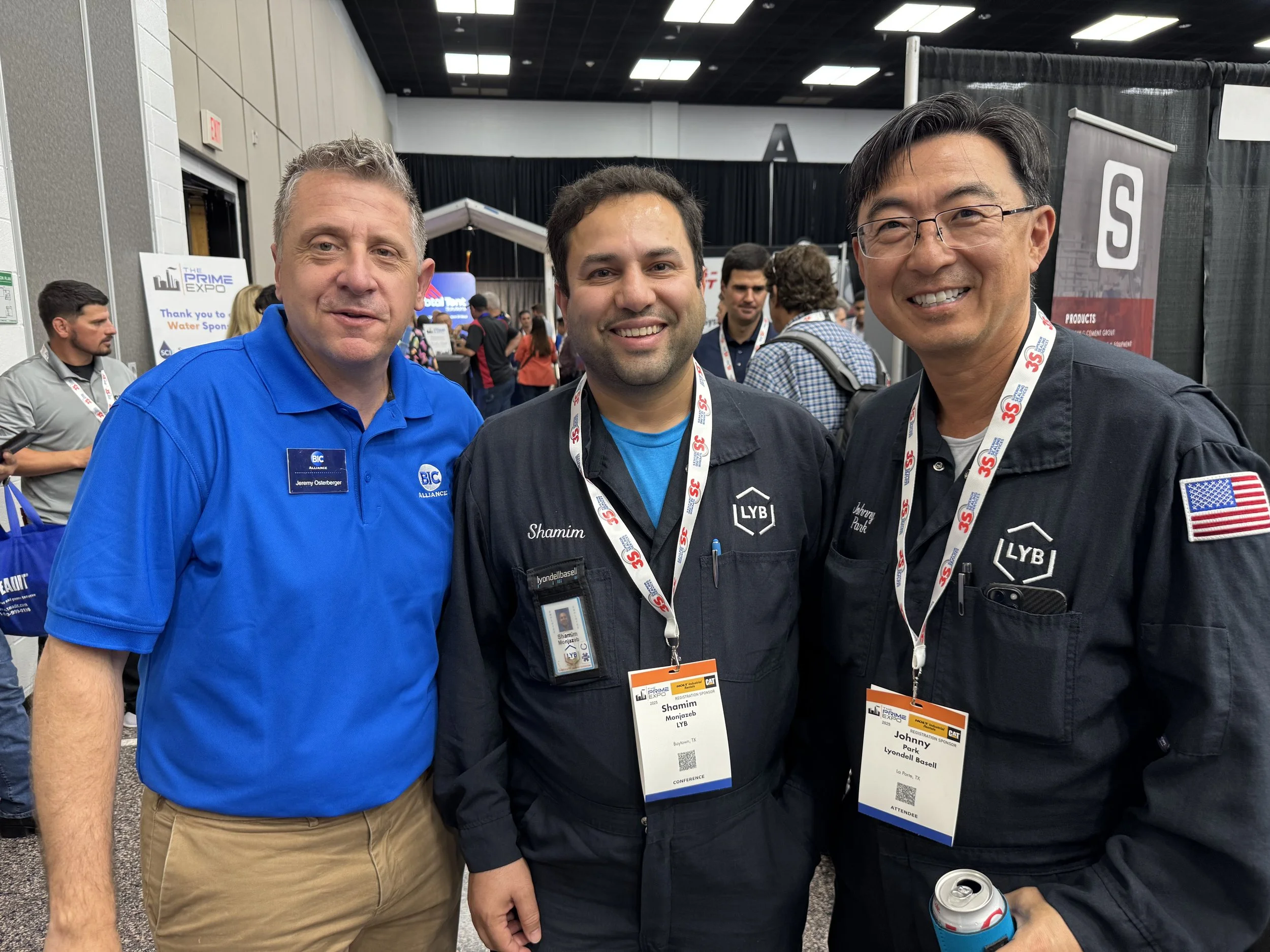 Three men standing together at a conference, smiling at the camera, wearing badges and black and blue uniforms. The background shows other attendees and exhibition booths.