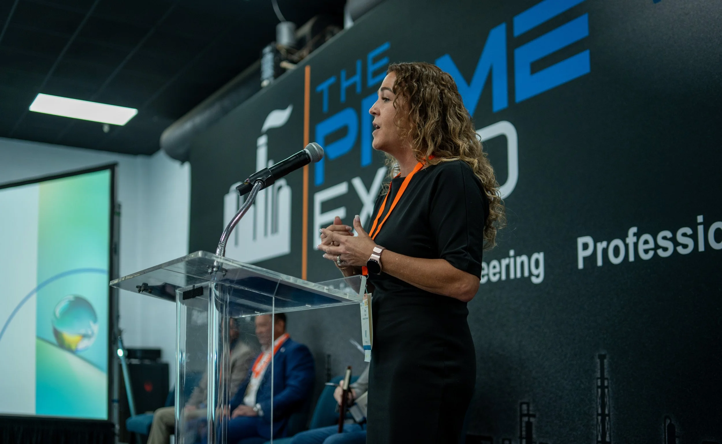 A woman speaking at a podium during a conference, with a backdrop displaying the words 'The Prime Expo' and part of a screen showing a computer desktop.
