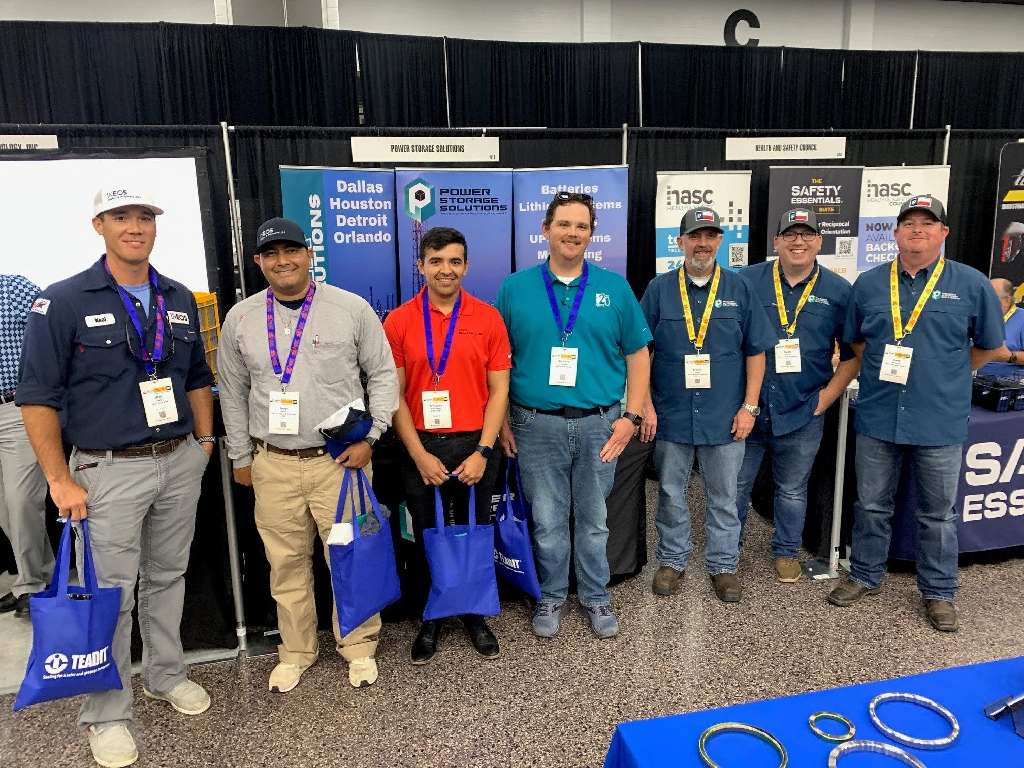 Group of seven men standing in front of a booth at a conference or trade show, wearing badges and holding blue tote bags, with banners and display stands behind them.