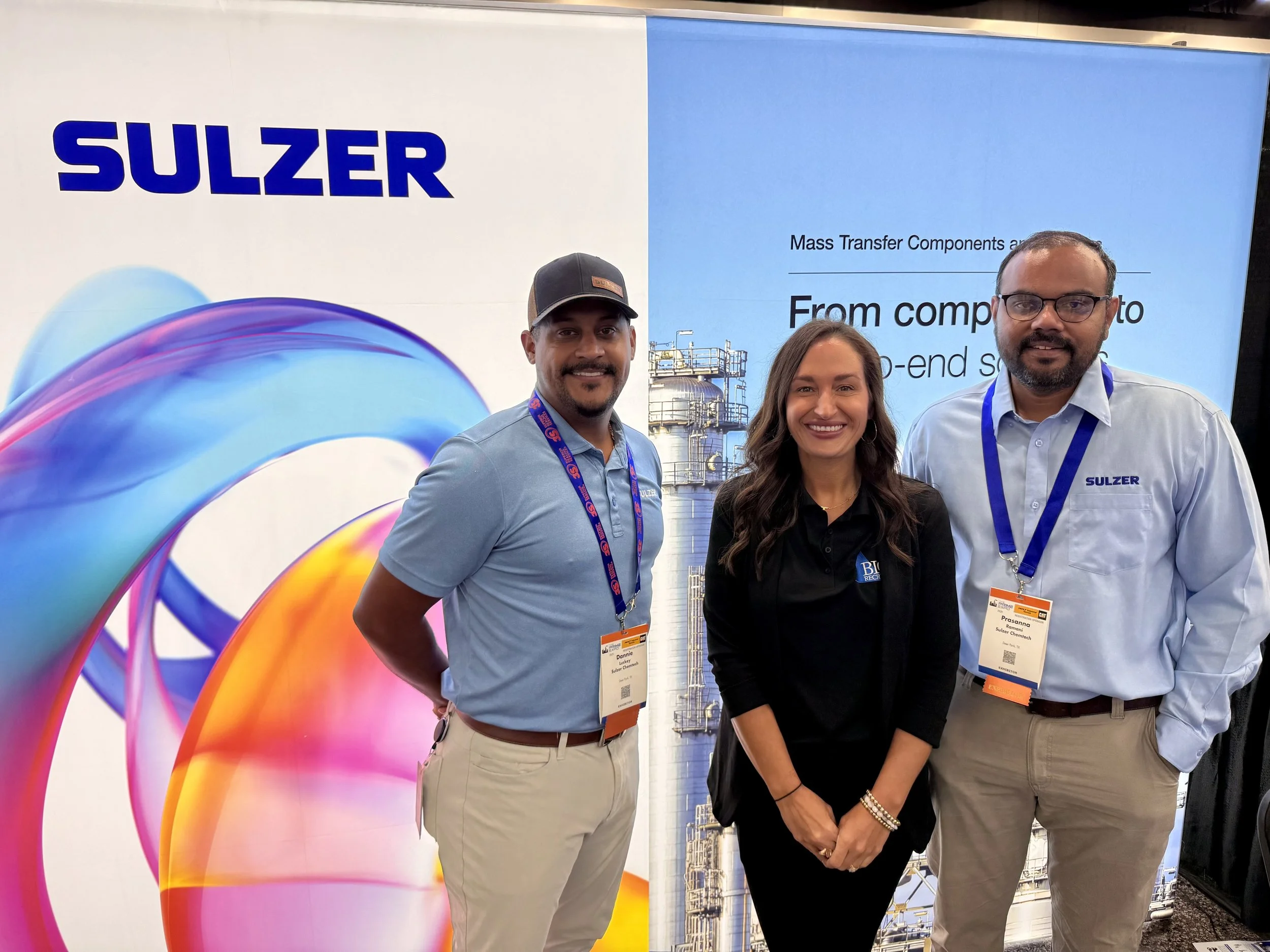 Three people standing in front of a colorful banner with the logo 'SULZER' and industrial equipment background, attending a professional event.