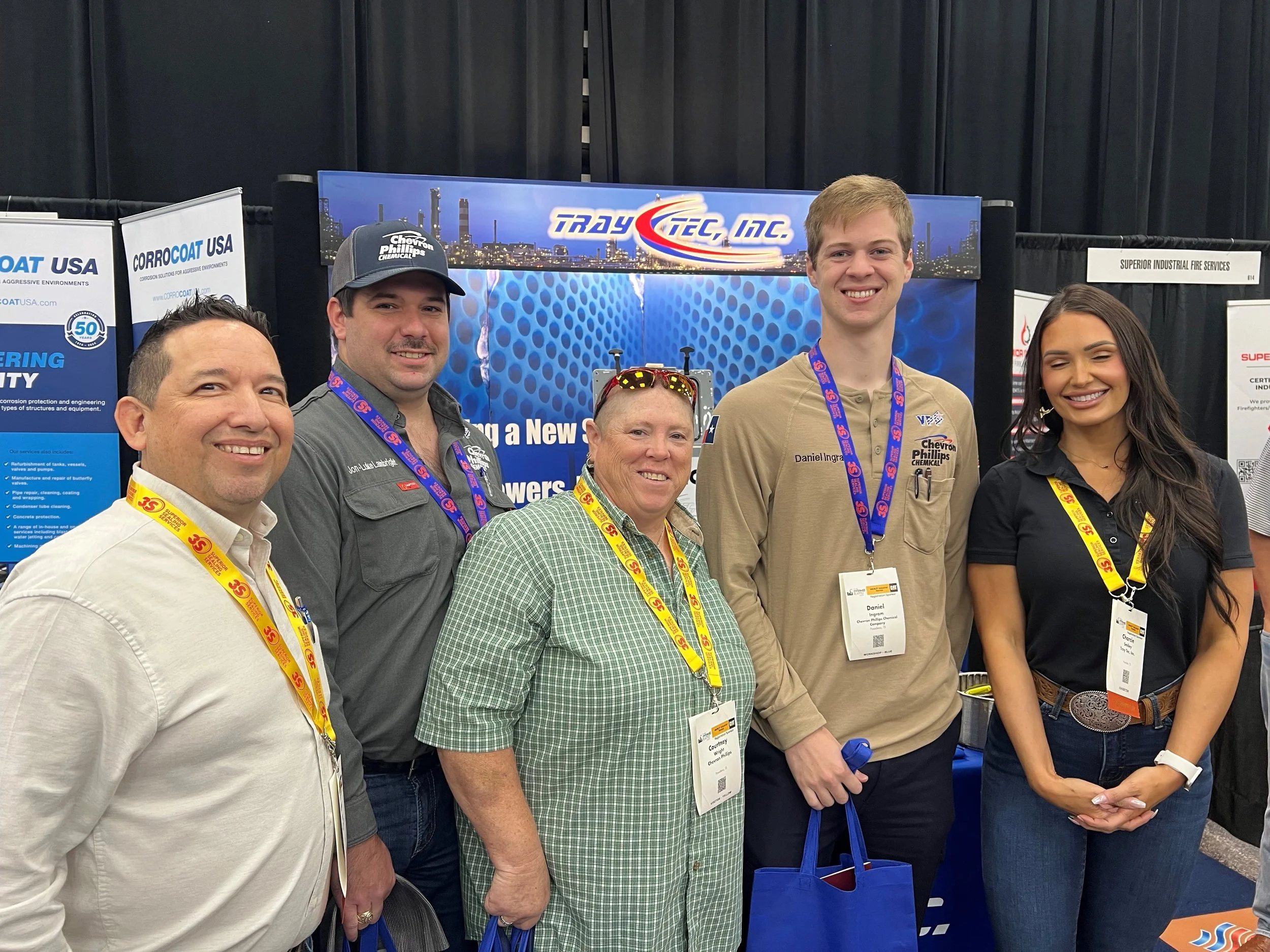 Group of five people standing in front of a booth at a conference, smiling, with blue lanyards and name tags.