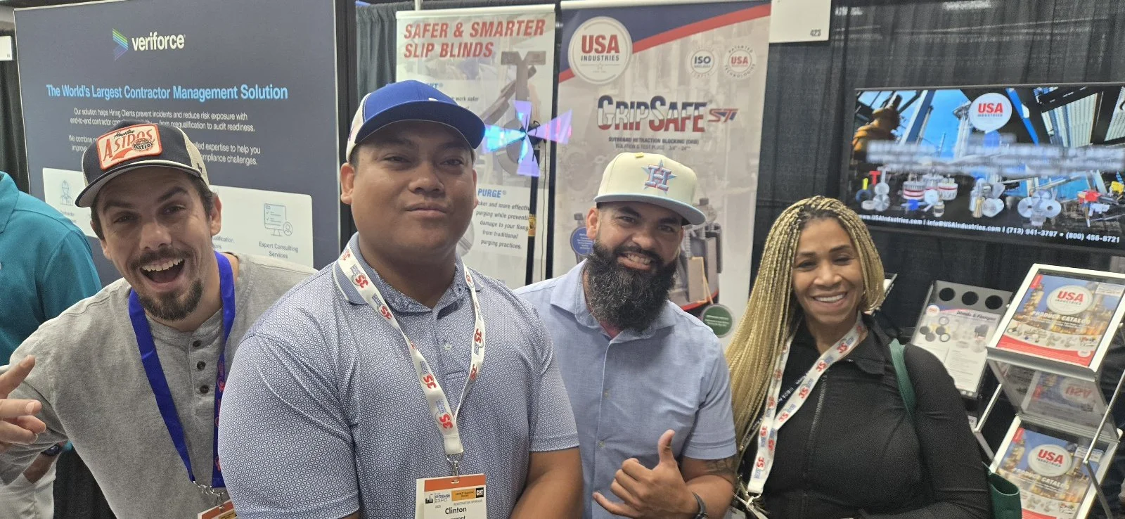 Four people smiling at a trade show booth with banners and screens displaying safety and construction equipment.