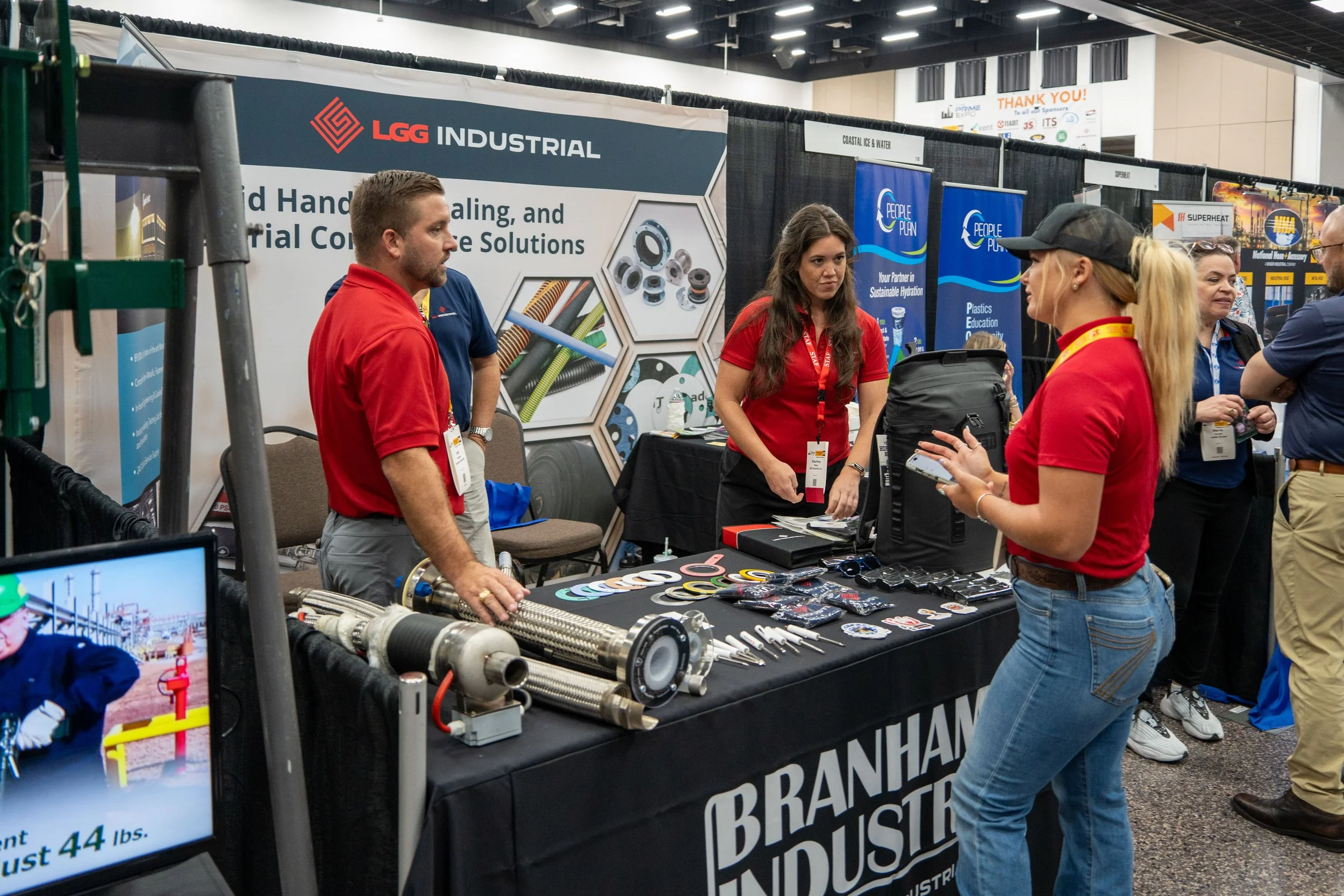 People at a trade show booth with branded signage for Branham Industries and LGG Industrial, displaying industrial equipment and promotional items, with attendees engaging in conversations.