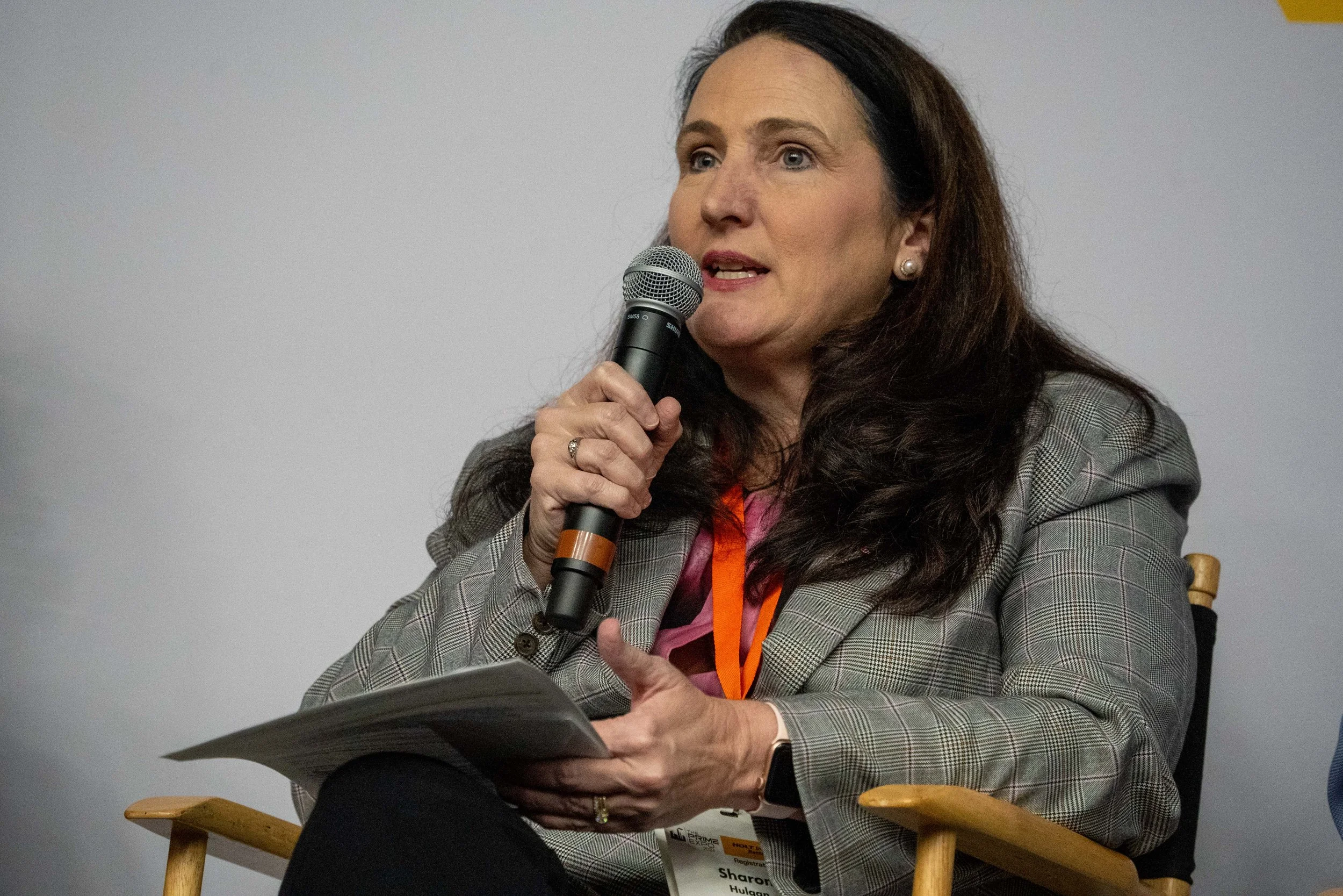 A woman with long dark hair, wearing a plaid blazer, is speaking into a microphone while seated in a wooden chair. She holds papers in her lap.