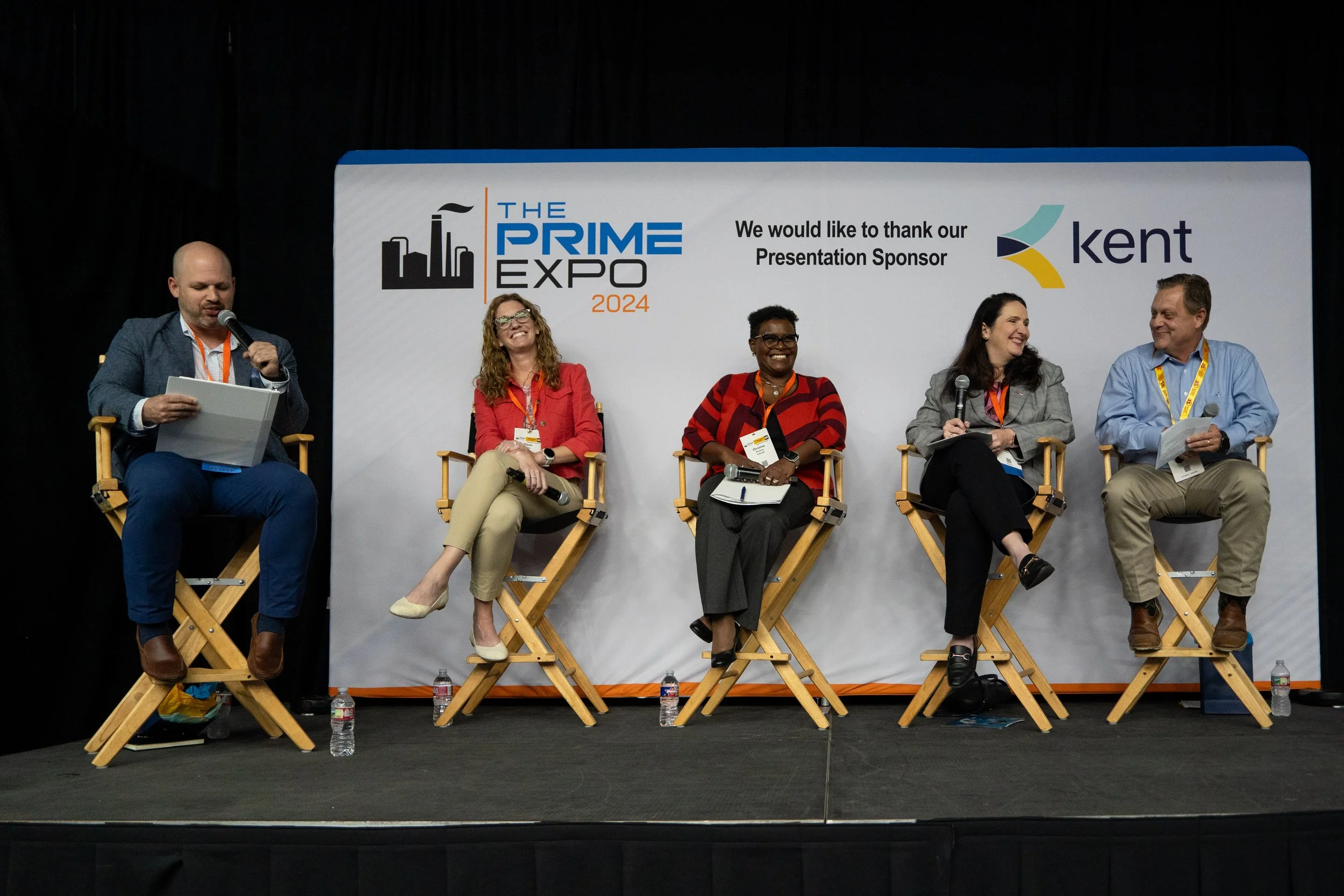 Five people sitting on stage in director's chairs during a panel discussion at The Prime Expo 2024, with a backdrop featuring the event logo and sponsor, Kent. One person on the far left is speaking into a microphone and holding papers; the others ar
