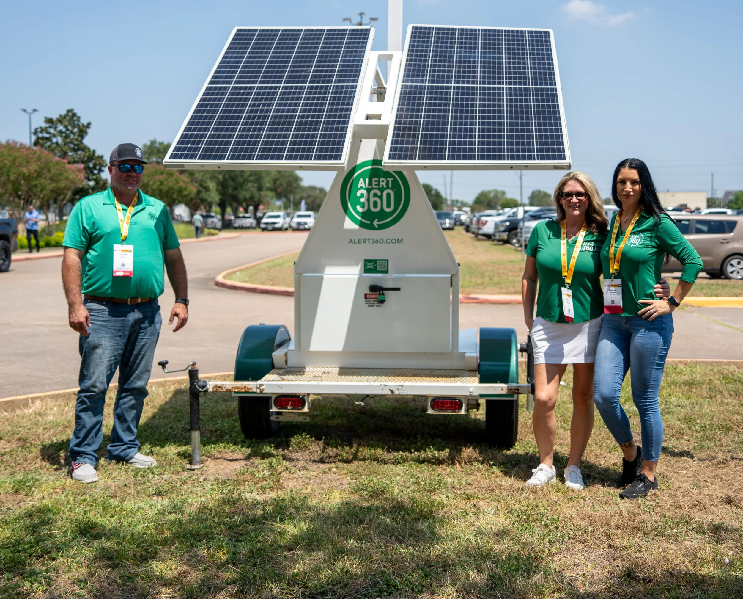 Three people standing outdoors in front of a mobile solar power generator with solar panels, on a sunny day, in a parking lot. Two women and one man, all wearing green shirts, are posing for the photo.