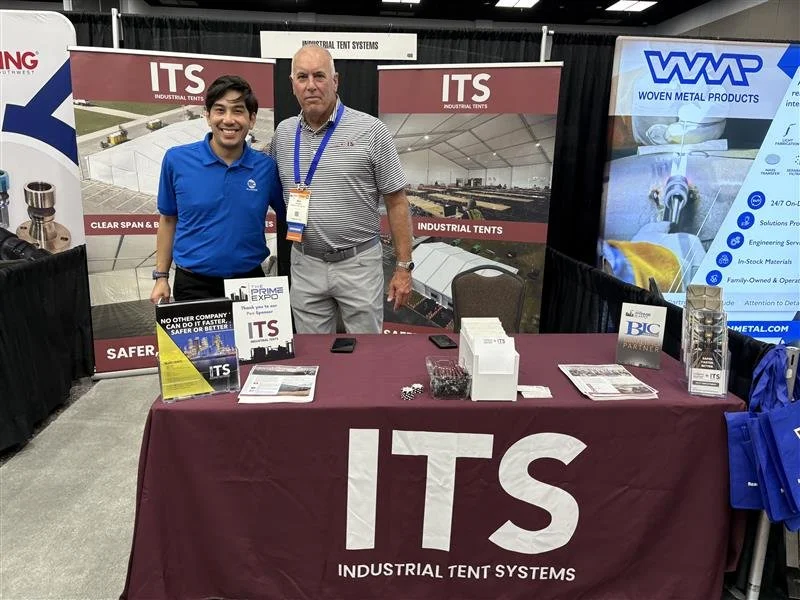 Two men standing behind a booth labeled 'ITS Industrial Tent Systems' at a trade show, with banners and promotional materials displayed. The man on the left is smiling and wearing a blue polo shirt, while the man on the right has a serious expression