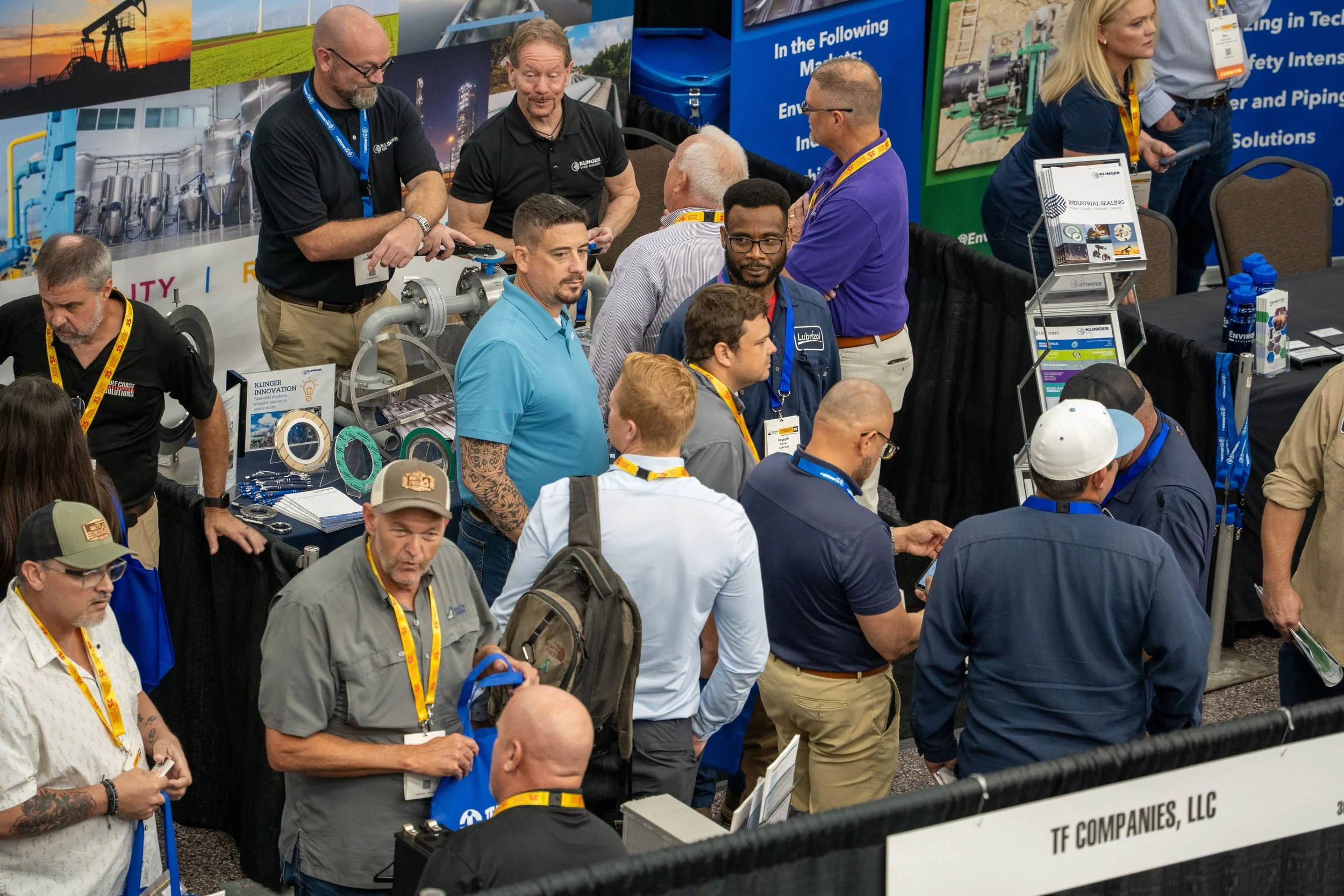 A crowded trade show booth with many people engaging and examining industrial equipment and products. The booth features displays, informational signs, and promotional materials related to industrial sealing solutions and other engineering products.