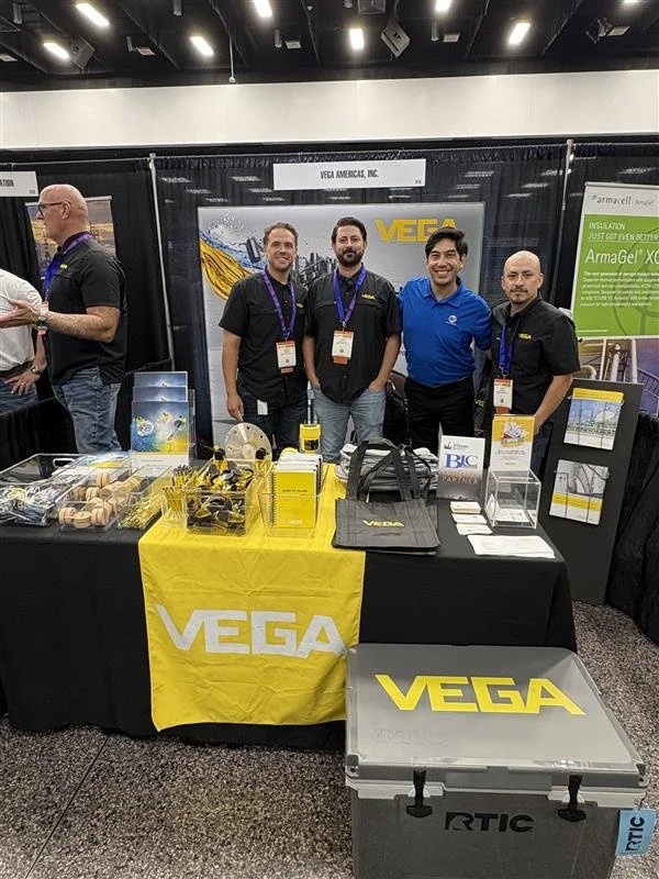 Four men standing behind a trade show booth for VEGA, displaying tools and equipment. The booth has a yellow and black color scheme with the VEGA logo on a table cover and cooler.