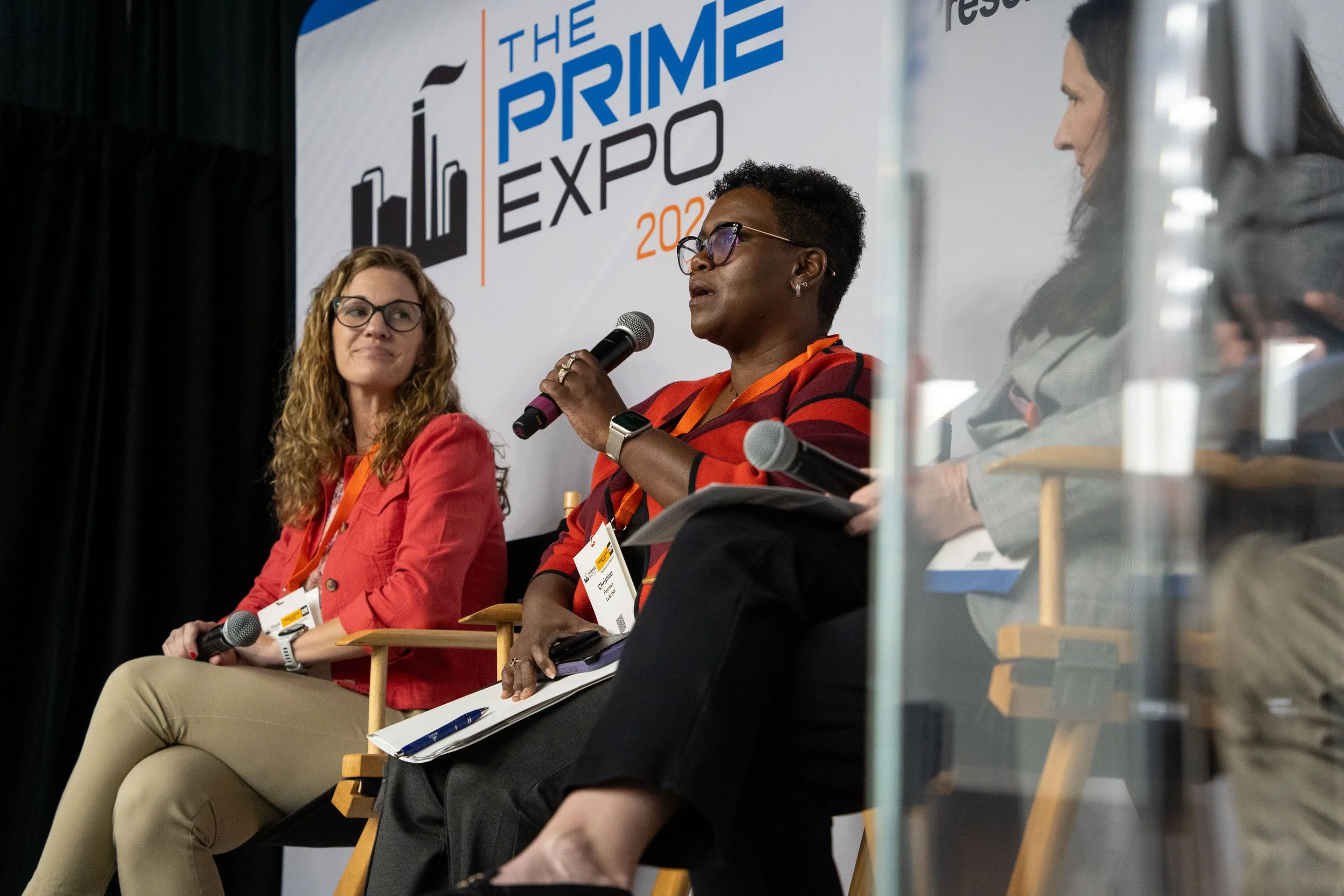 Three women sitting on stage at The Prime Expo 2023, participating in a panel discussion, with one woman speaking into a microphone, and a banner for the event visible in the background.